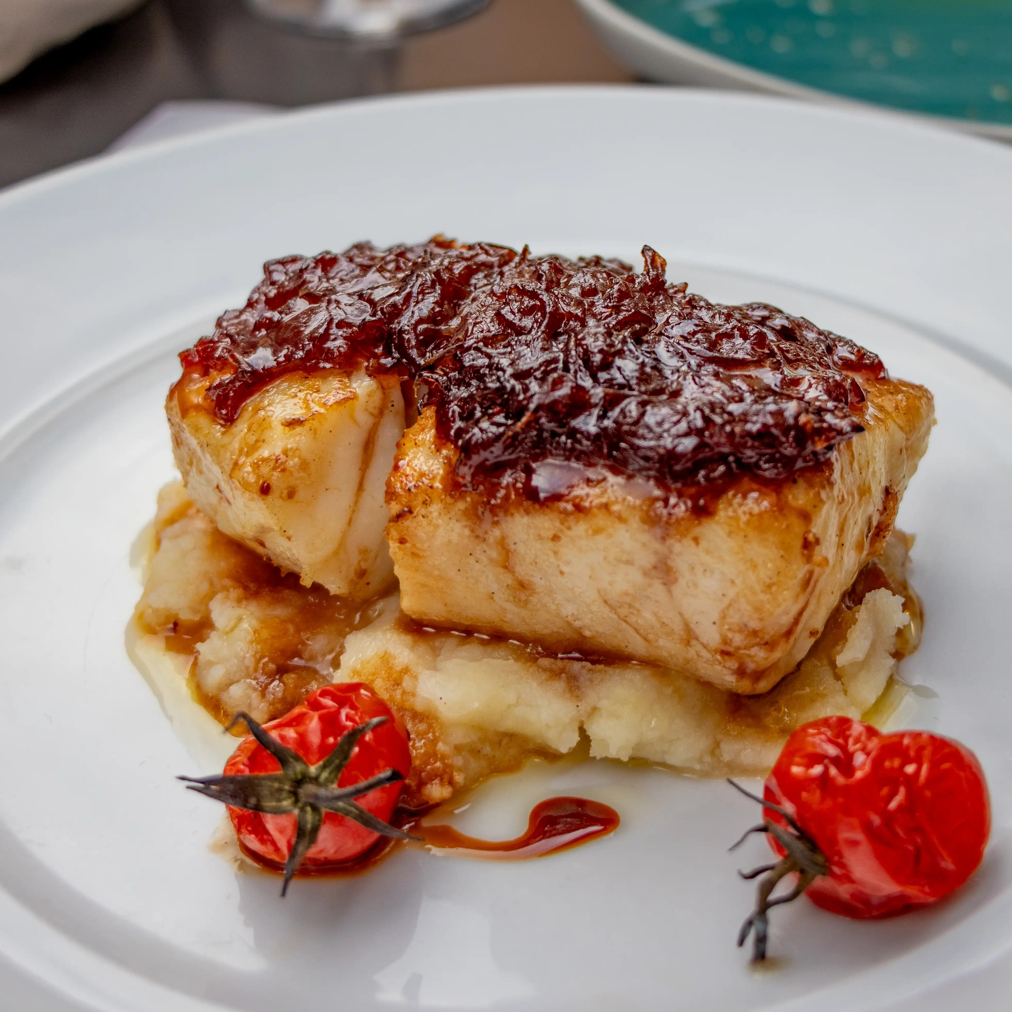 A plated piece of fish topped with caramelized onions, served over mashed potatoes with two small tomatoes on the side.