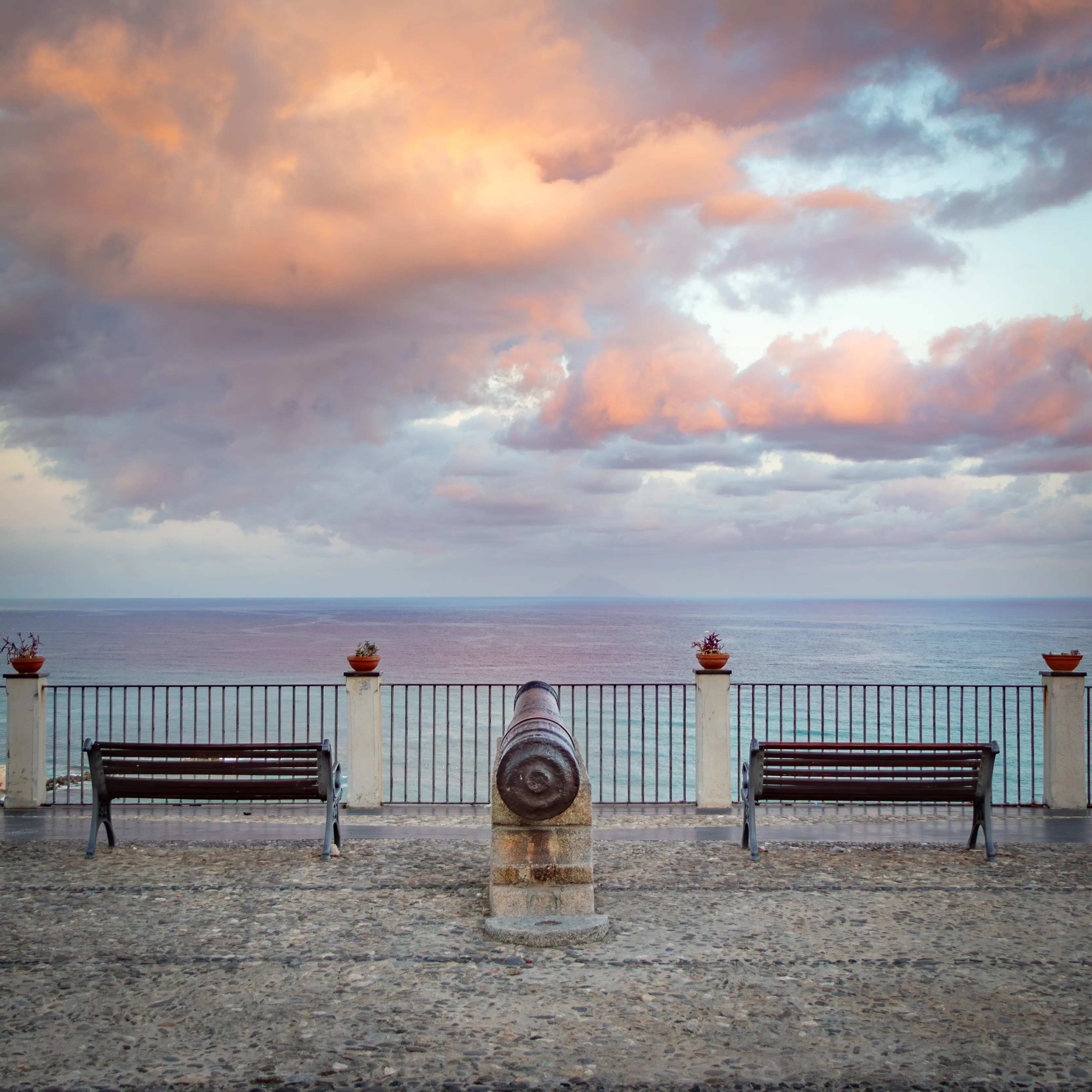 Cannon mounted on a stone base facing the sea with benches and a railing behind it.