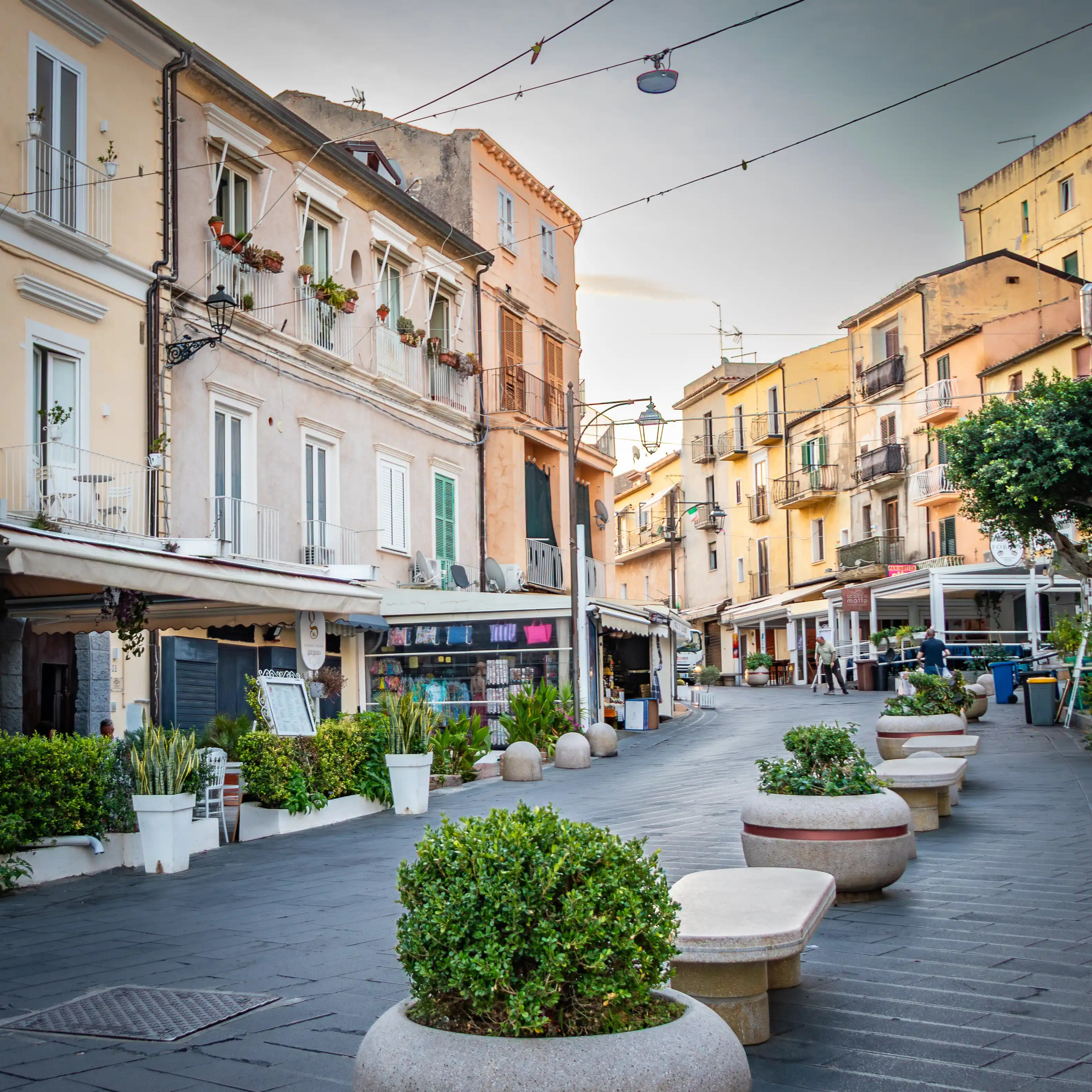 Pedestrian street lined with buildings, shops, and large round planters with greenery.