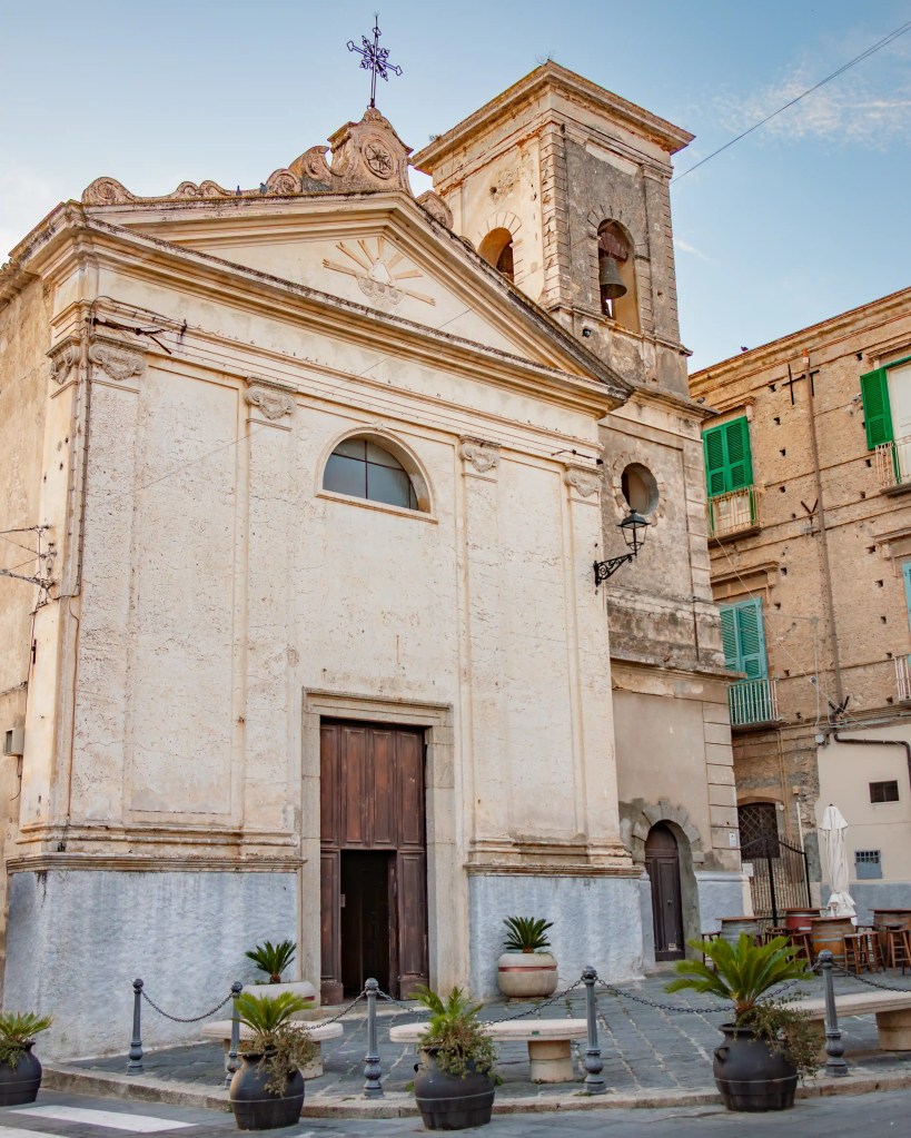 Church façade with triangular pediment, bell tower, and wooden doors facing a small square.