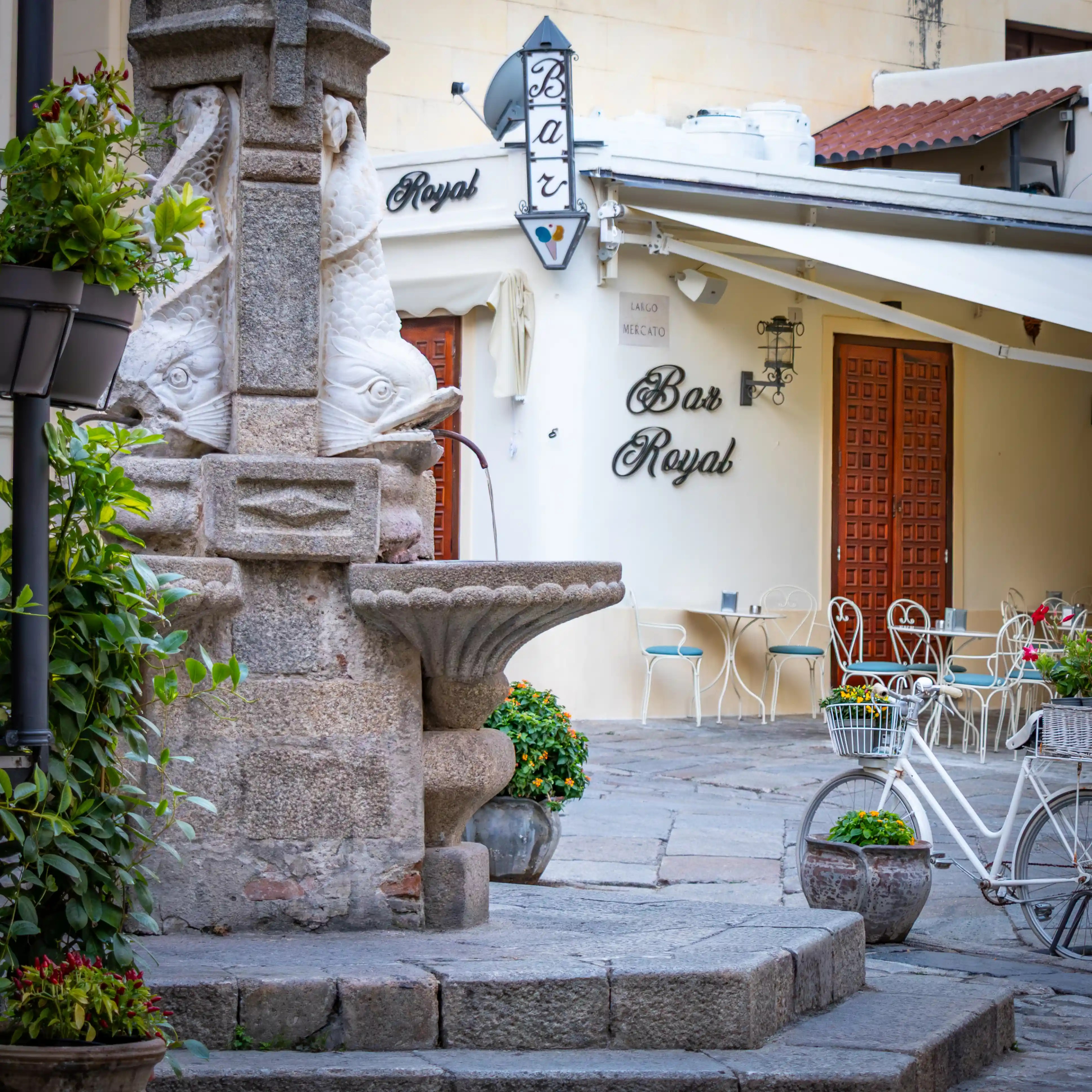 Stone fountain with carved fish figures in the foreground and a building with a “Bar Royal” sign and outdoor seating behind it.