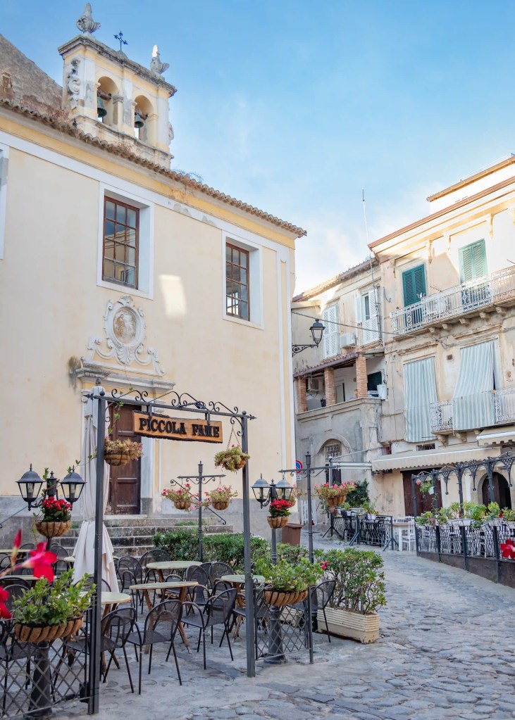 Church façade with a small bell tower and a sign reading “Piccola Fame” above outdoor seating in a square.