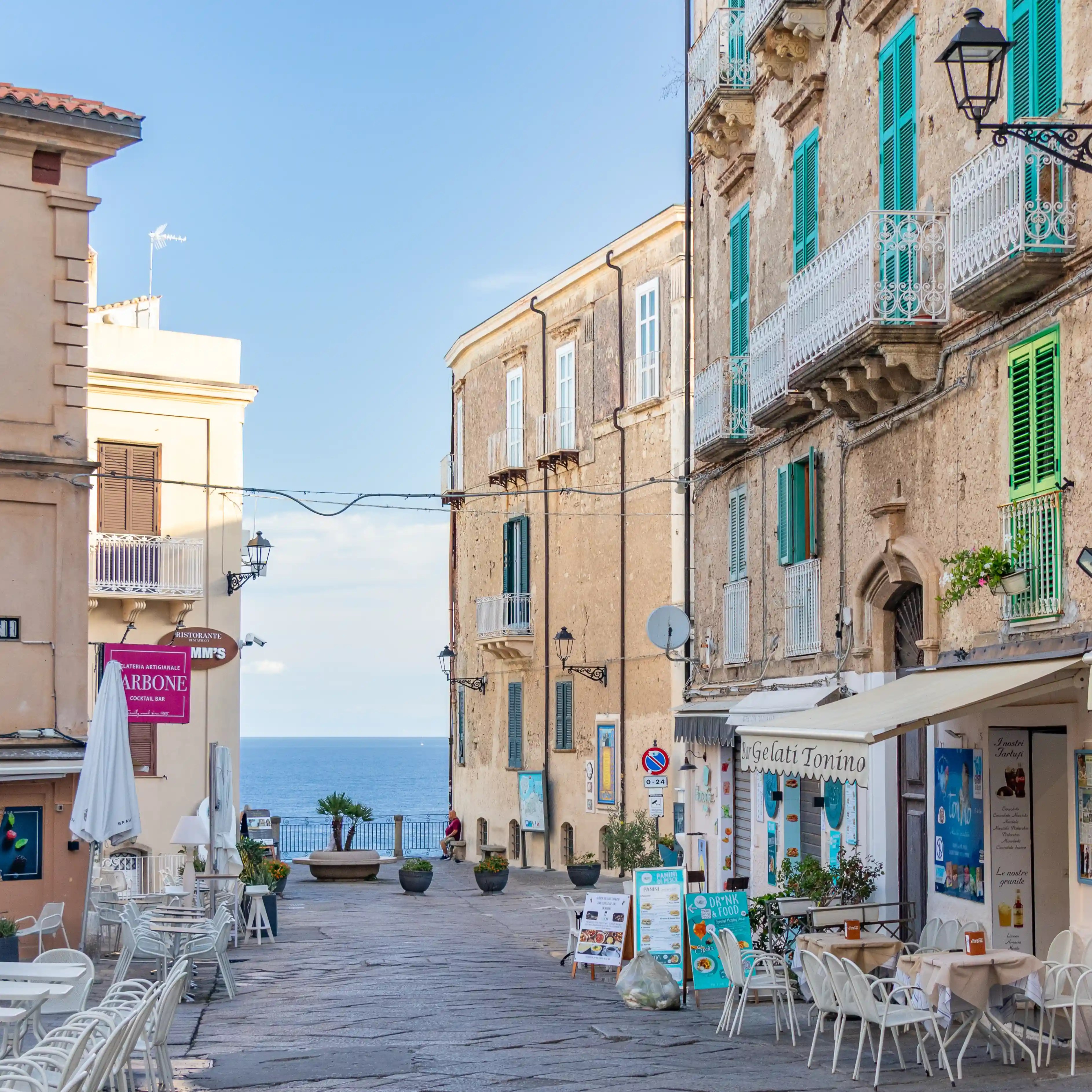 Street lined with buildings, shops, and outdoor seating leading toward the sea in the distance.