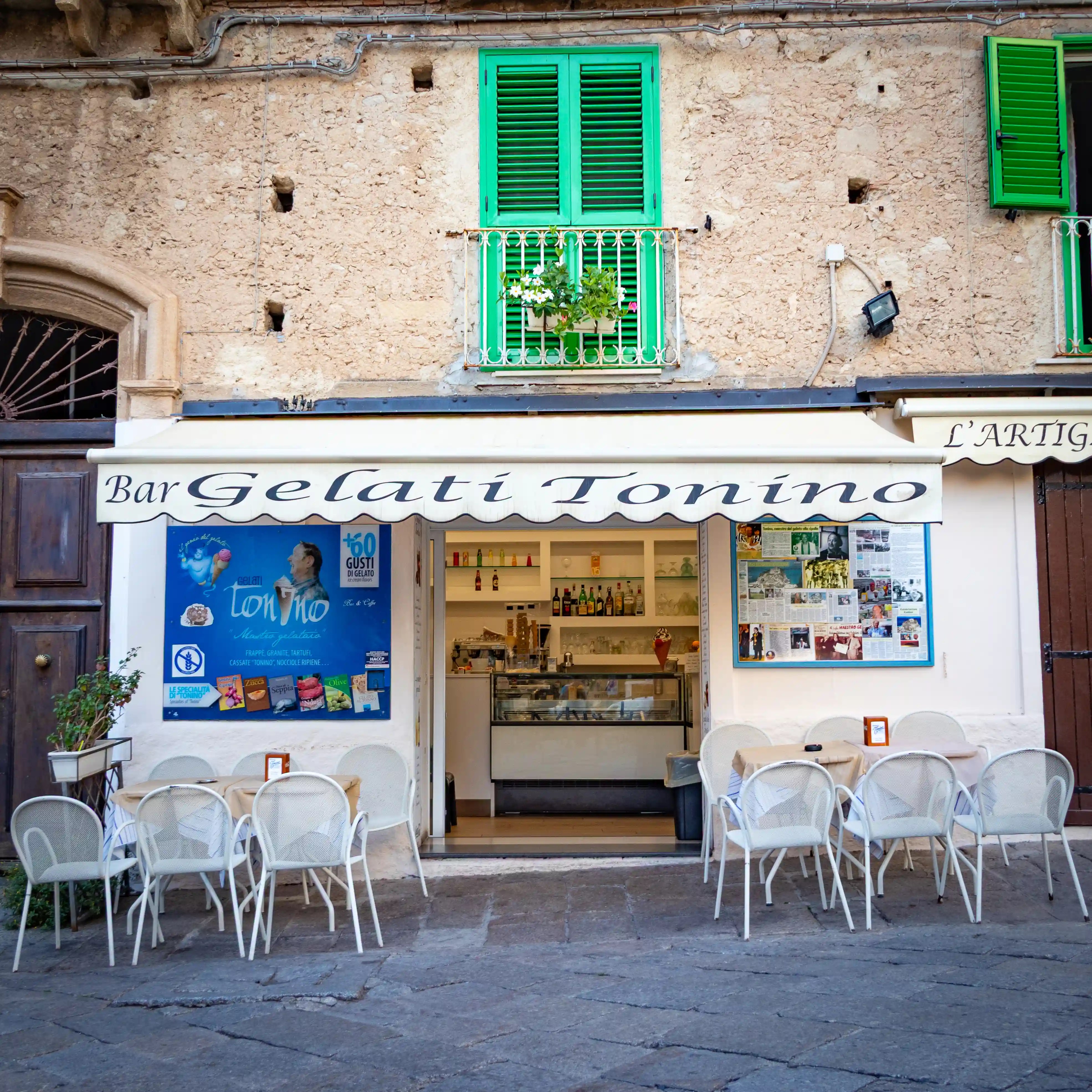 Storefront with sign reading “Bar Gelati Tonino” with outdoor seating and a display counter inside.