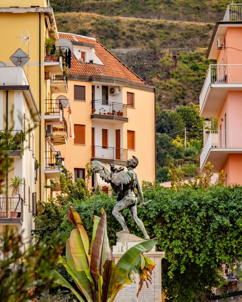 Statue of a winged figure on a pedestal surrounded by plants with residential buildings and hills in the background.