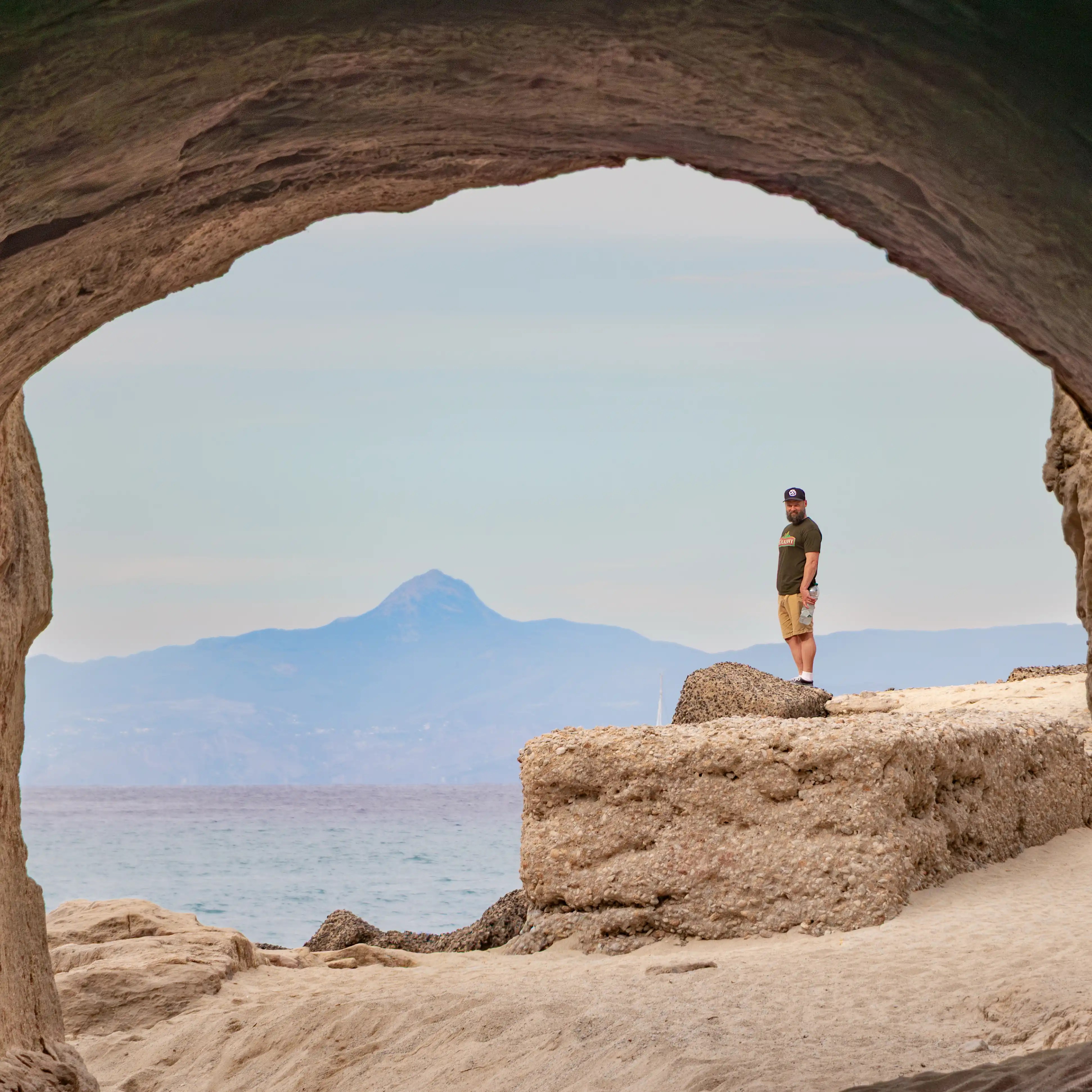 A person standing on a rocky ledge framed by a natural stone arch with the sea and distant mountains behind.