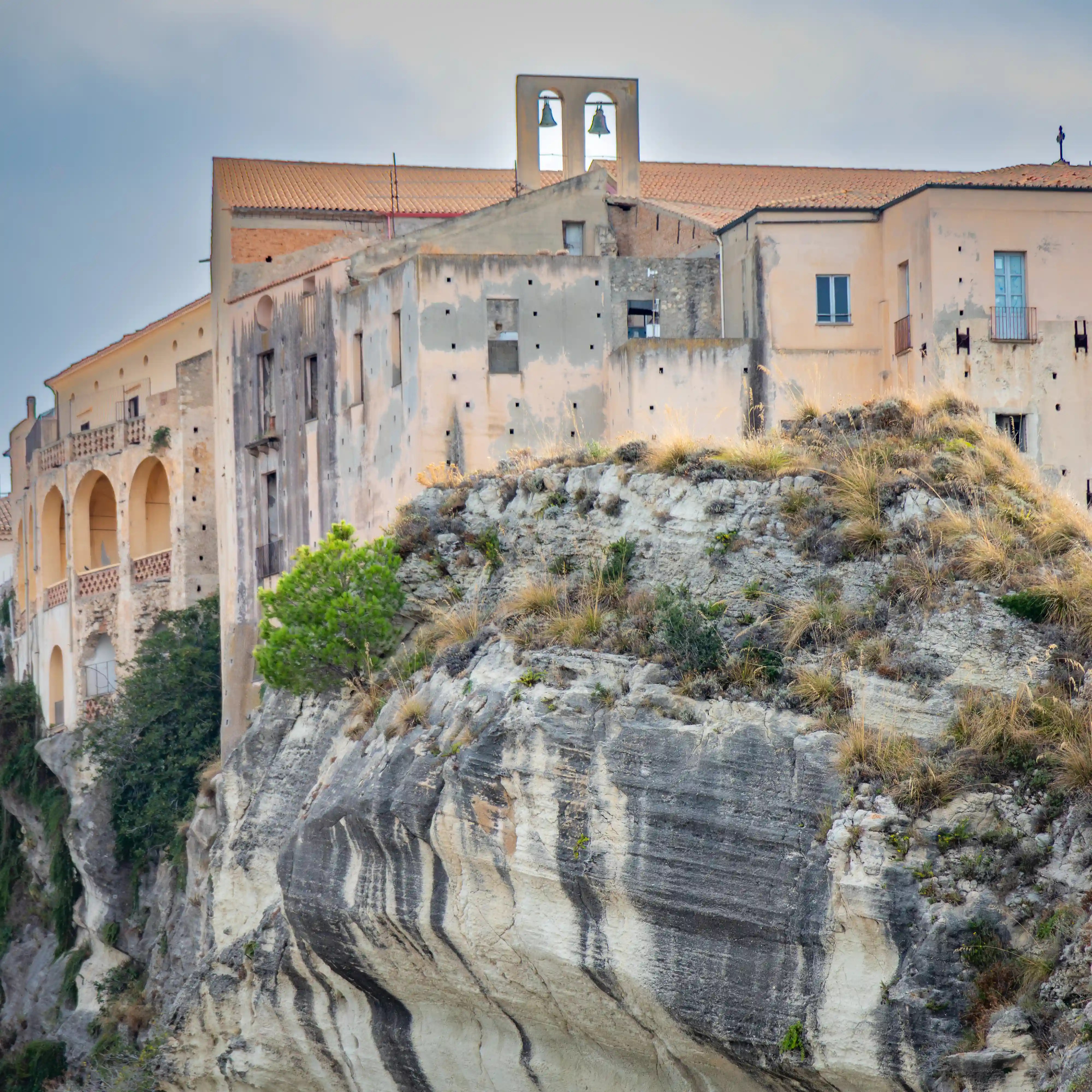 Weathered building with arched balconies and a small bell structure sitting on a rocky cliff.