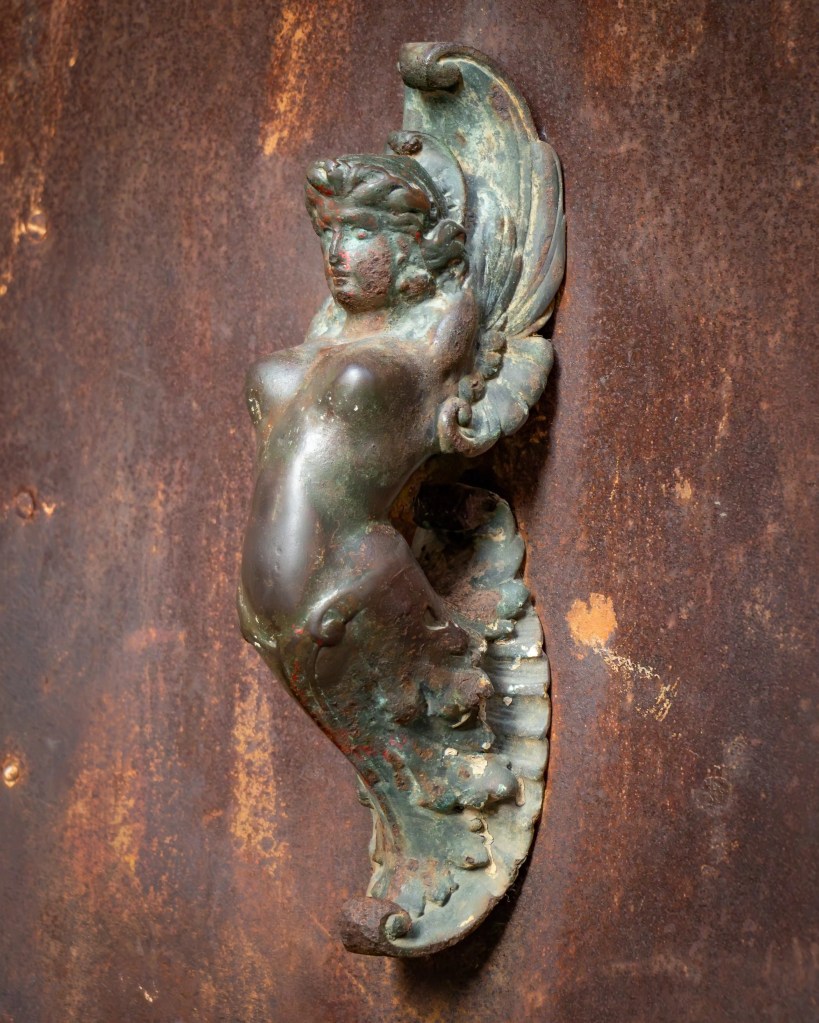 Metal door knocker shaped like a winged figure mounted on a weathered brown door.