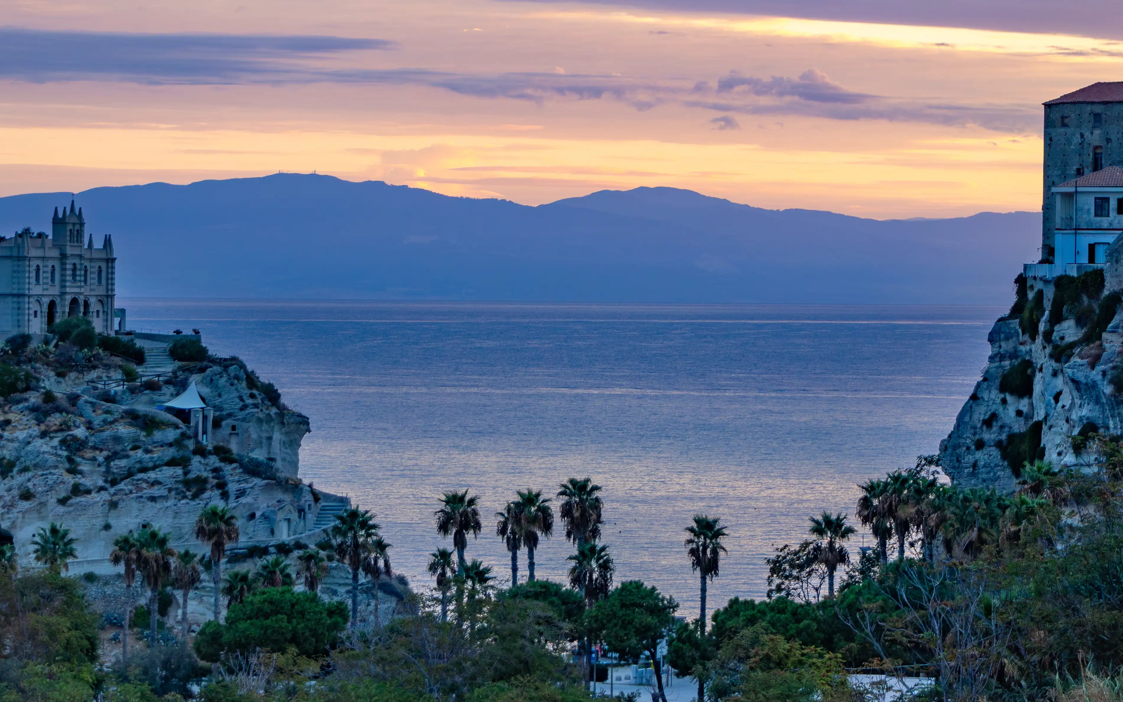 Sunrise over the sea framed by two rocky cliffs, with palm trees below and buildings along the cliff edges.