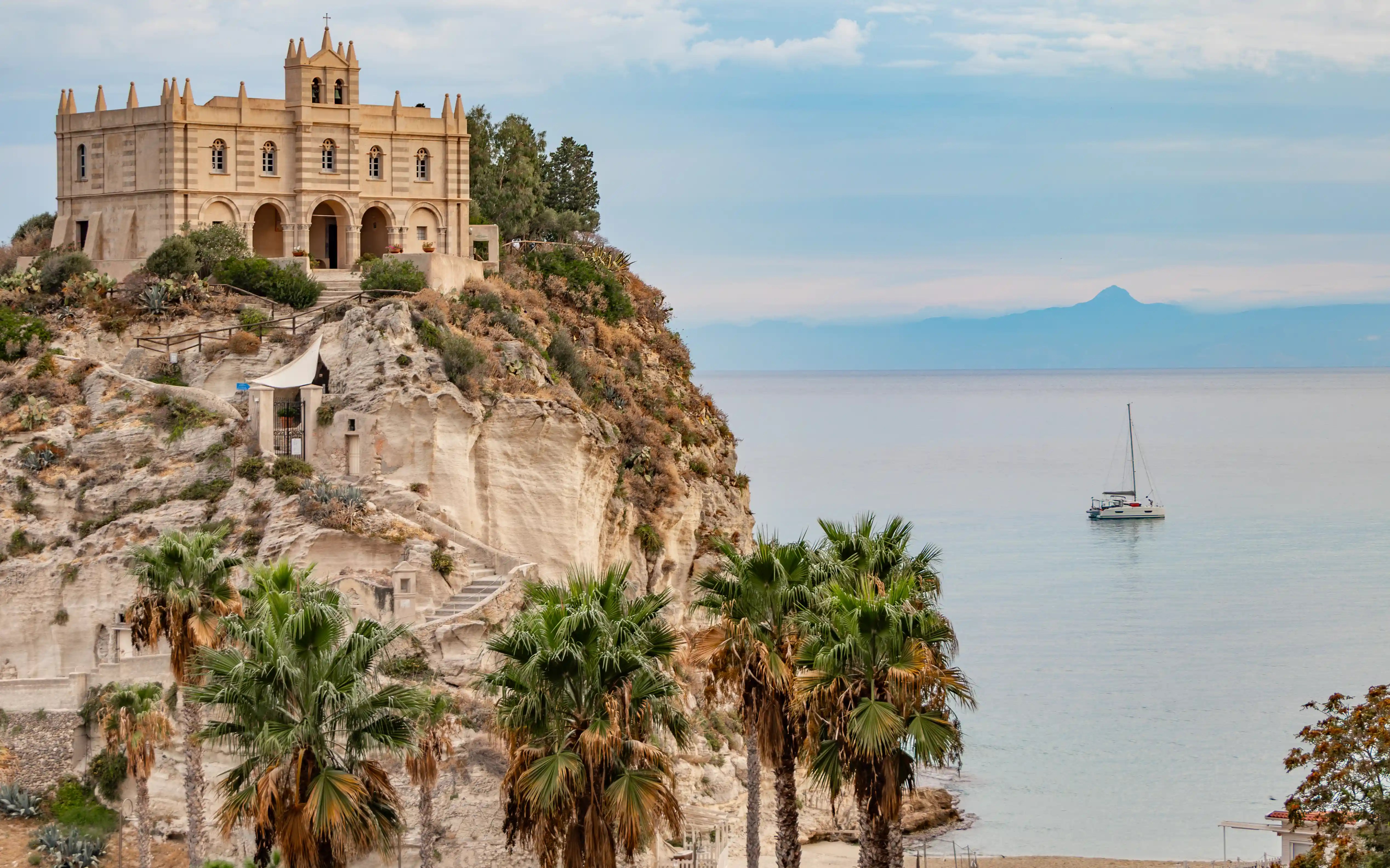 Church on a rocky cliff above the sea with palm trees in the foreground and a sailboat in the water.