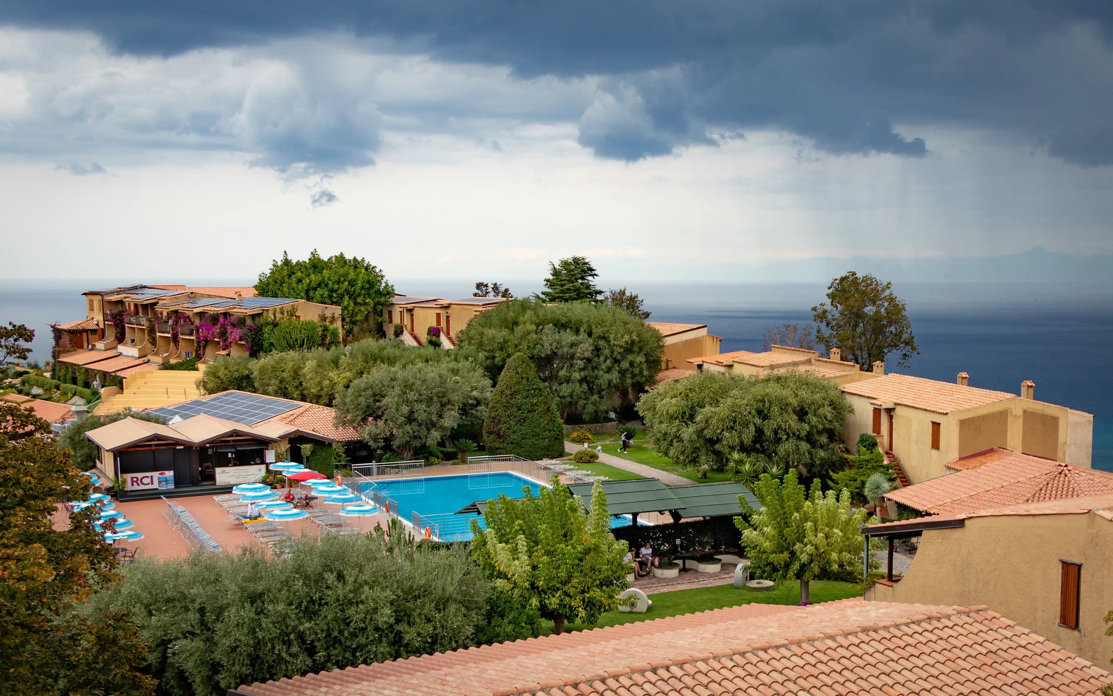 Resort area with a swimming pool, lounge chairs, and buildings overlooking the sea under cloudy skies.