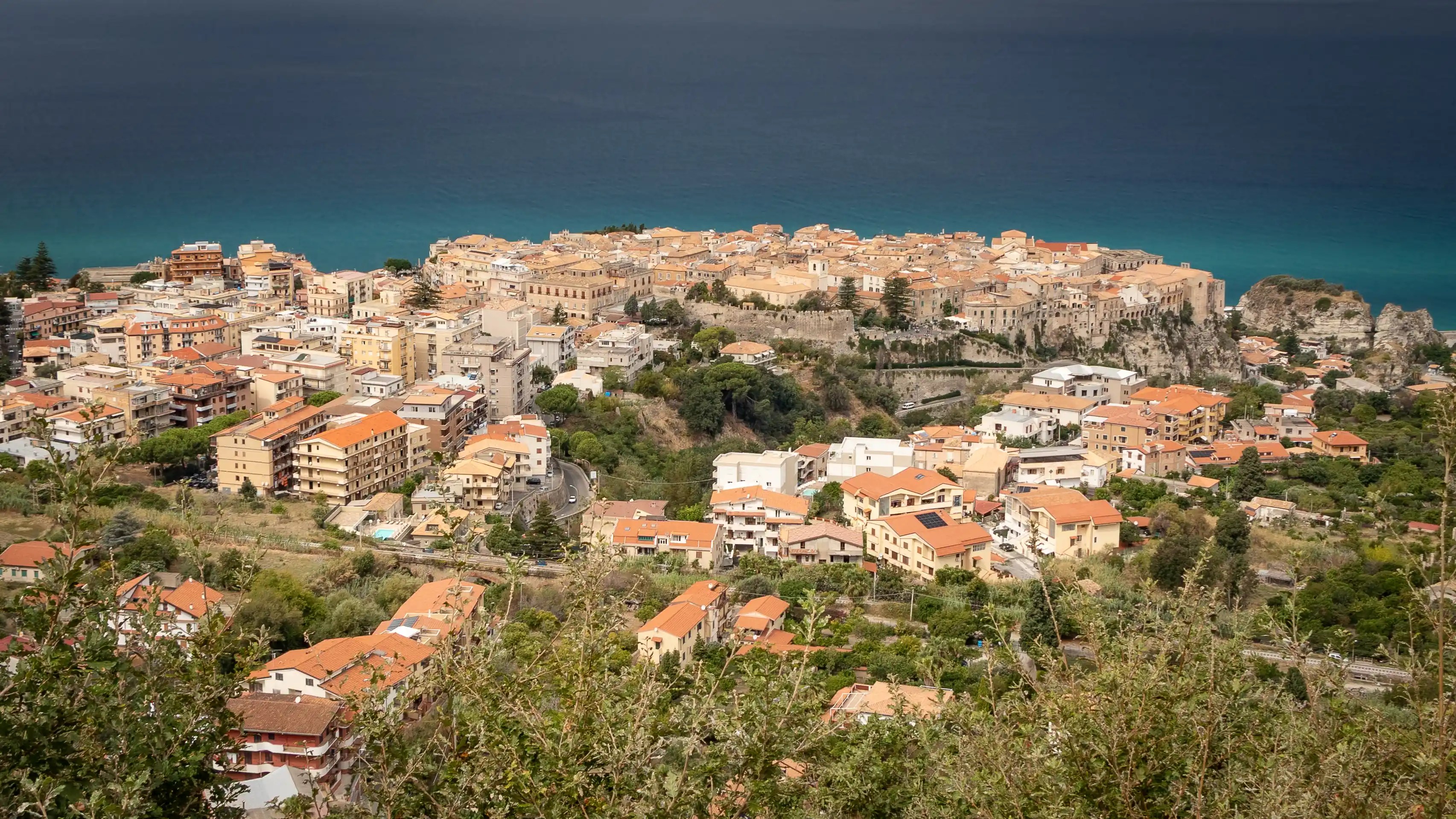 An elevated view of Tropea showing the historic center on a cliff above the sea with surrounding residential buildings in the foreground.