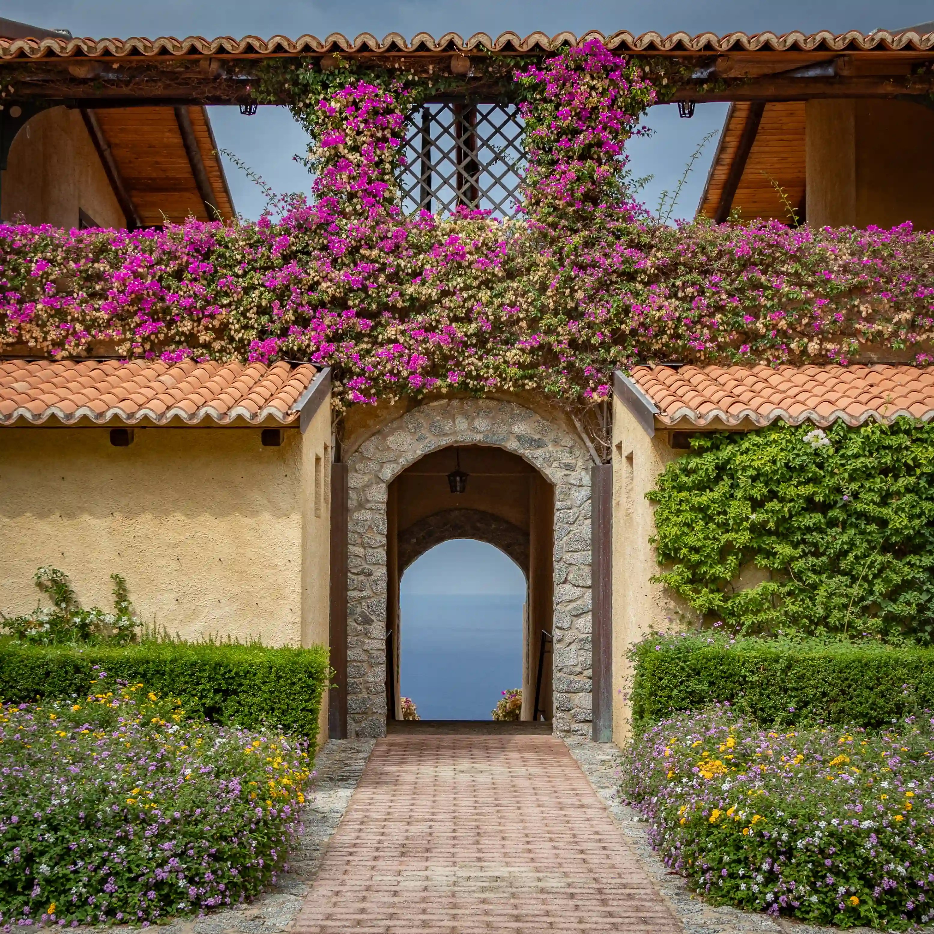 Stone archway framed by flowering vines and greenery leading to a view of the sea beyond.