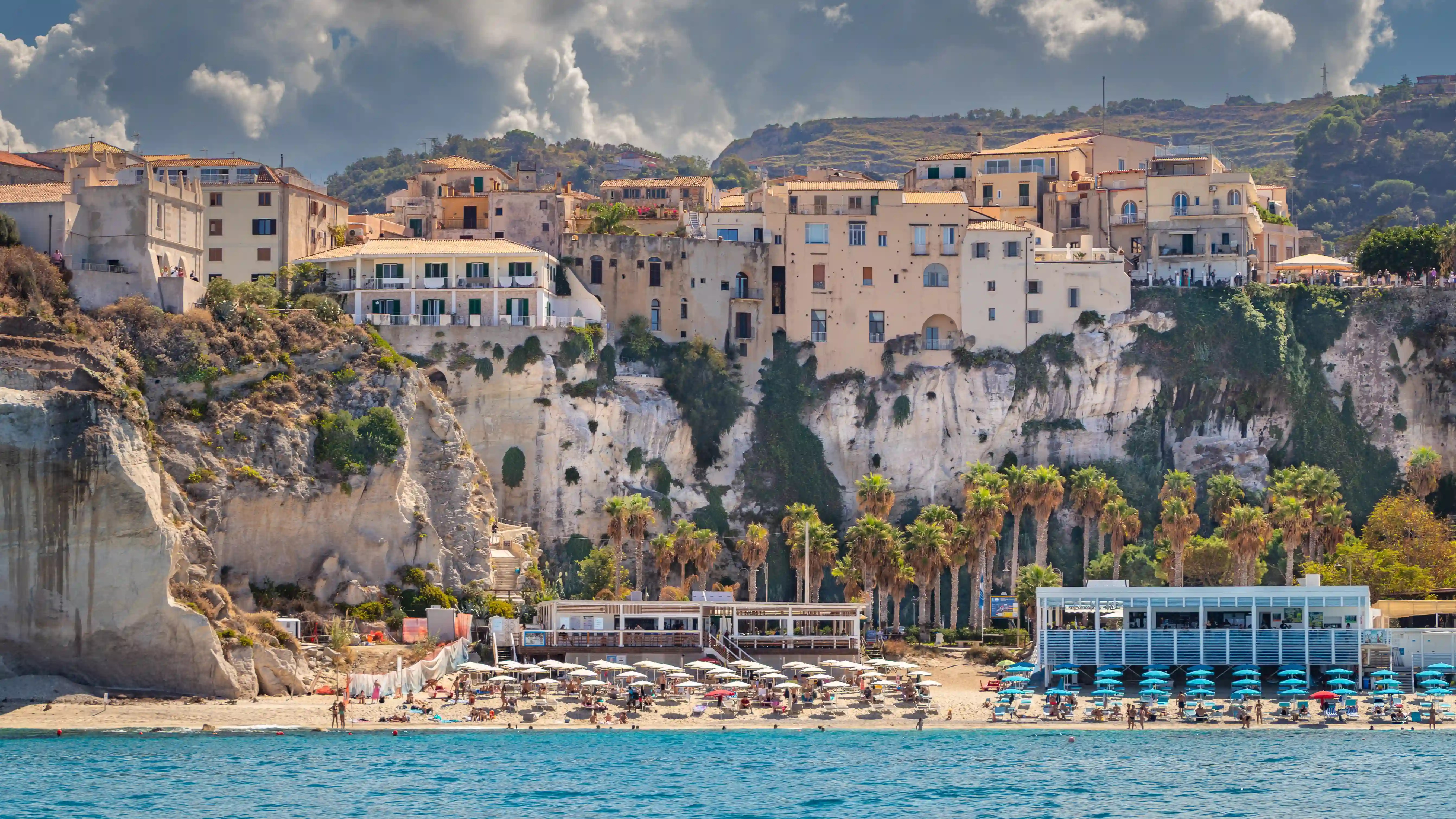 Buildings of Tropea’s old town sitting along steep cliffs above a beach and palm trees.