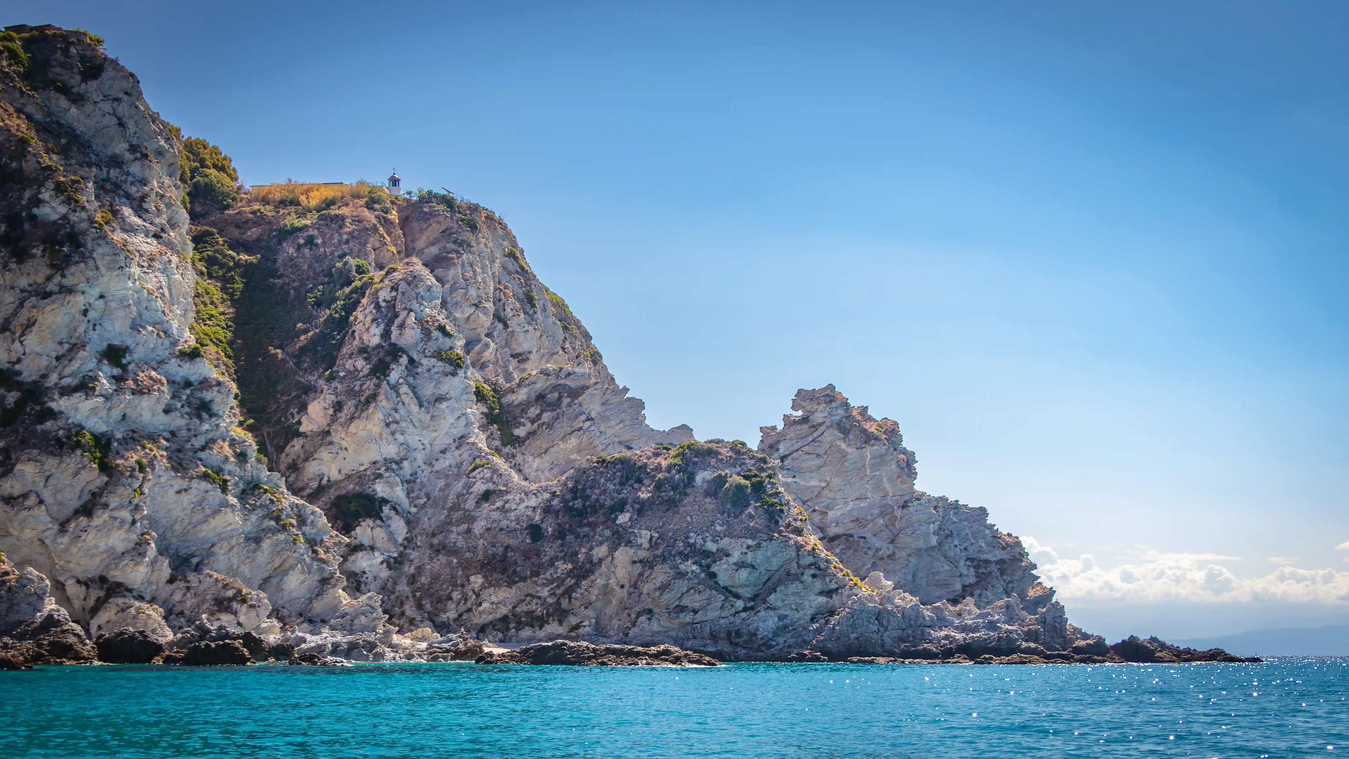 A rocky coastal cliff rising above turquoise water with a small lighthouse visible at the top.