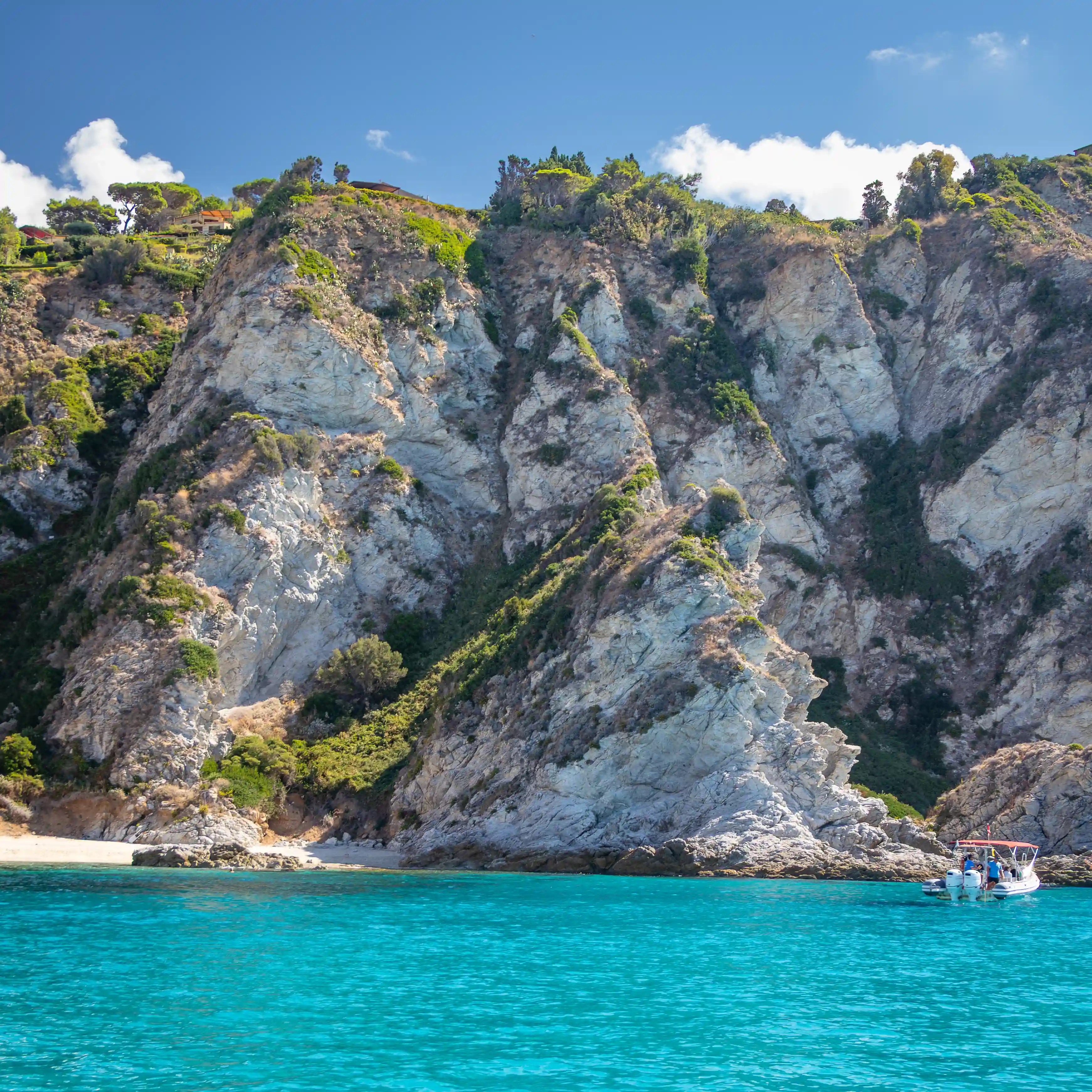A small boat floating near steep rocky cliffs above a narrow strip of beach and turquoise water.