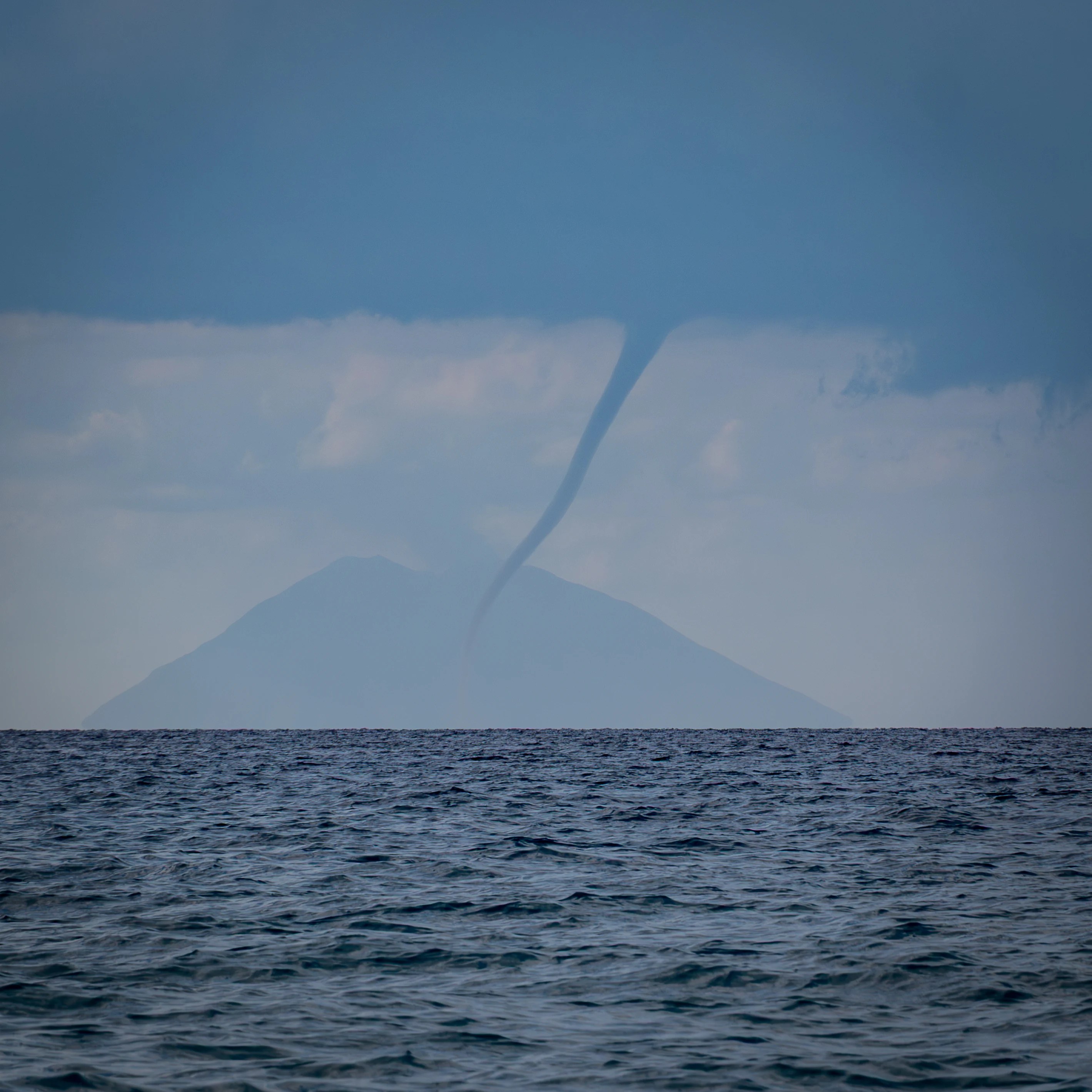 A waterspout extending from dark clouds down to the sea with a faint island silhouette in the background.