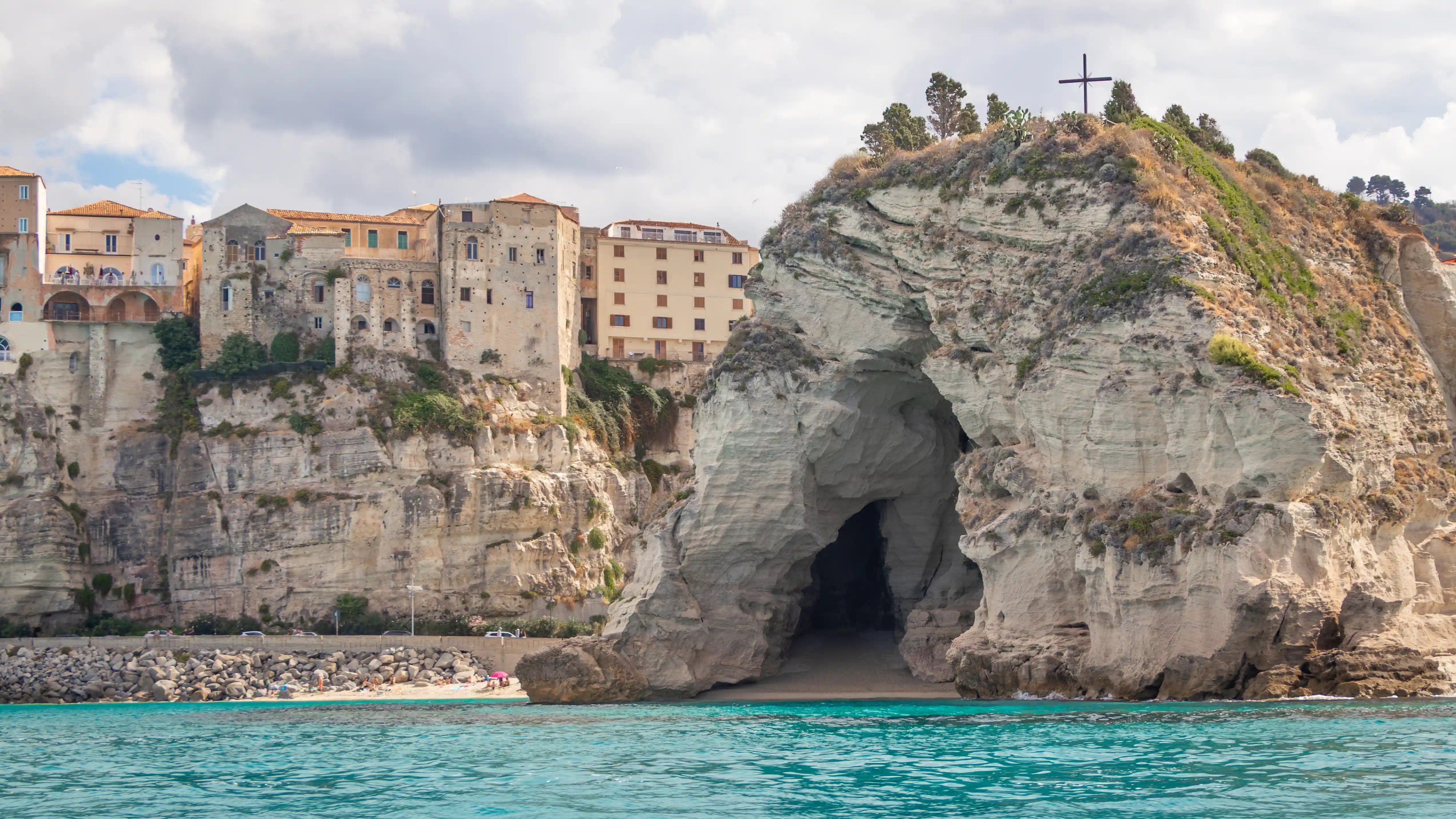 A large cave opening in the rock of L’Isola in Tropea with a cross on top and the old town cliffs in the background.