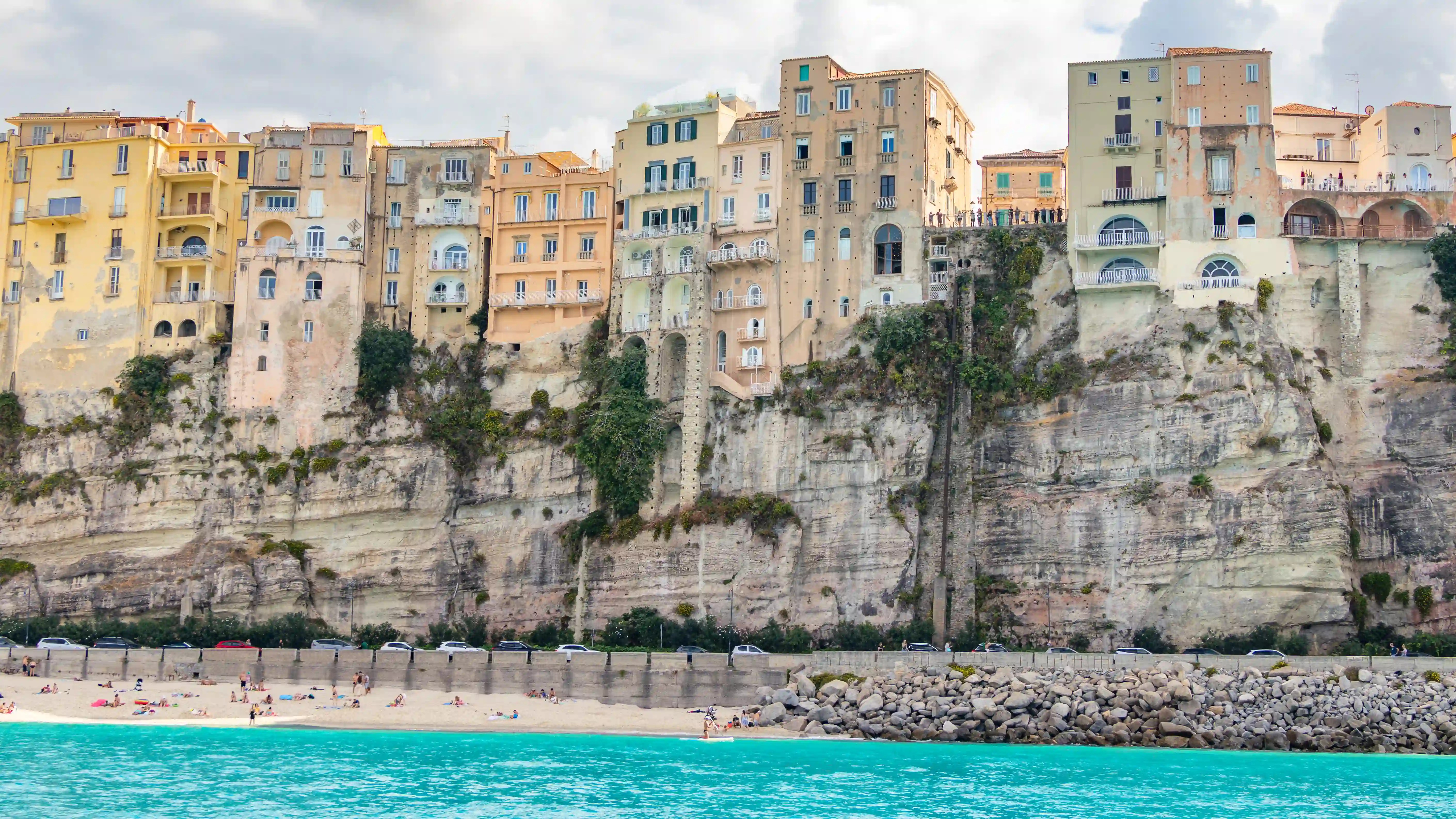 A view from the beach showing tall buildings in Tropea rising above a steep cliff with the shoreline below.