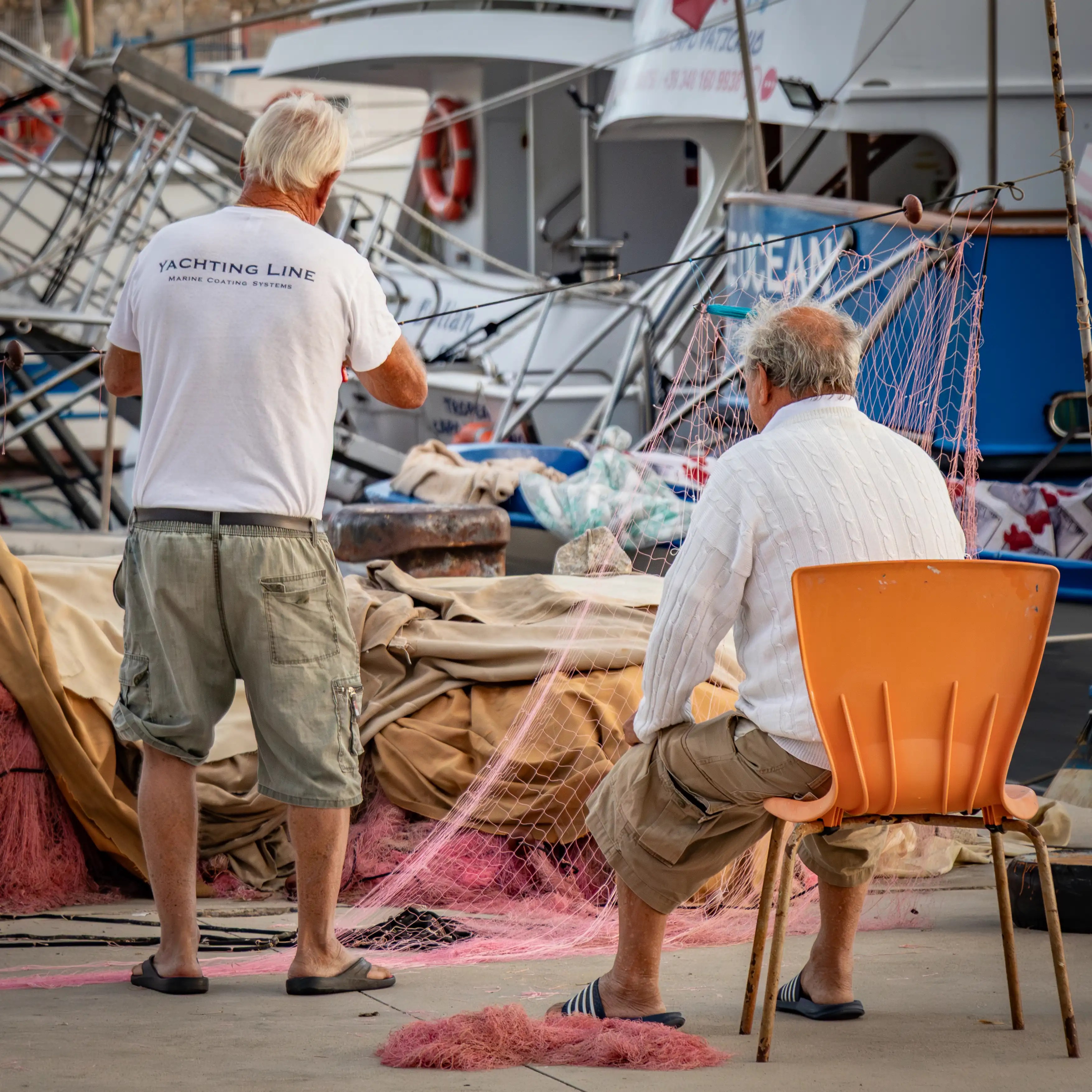 Two men working with fishing nets beside boats in a marina, one standing and one seated.