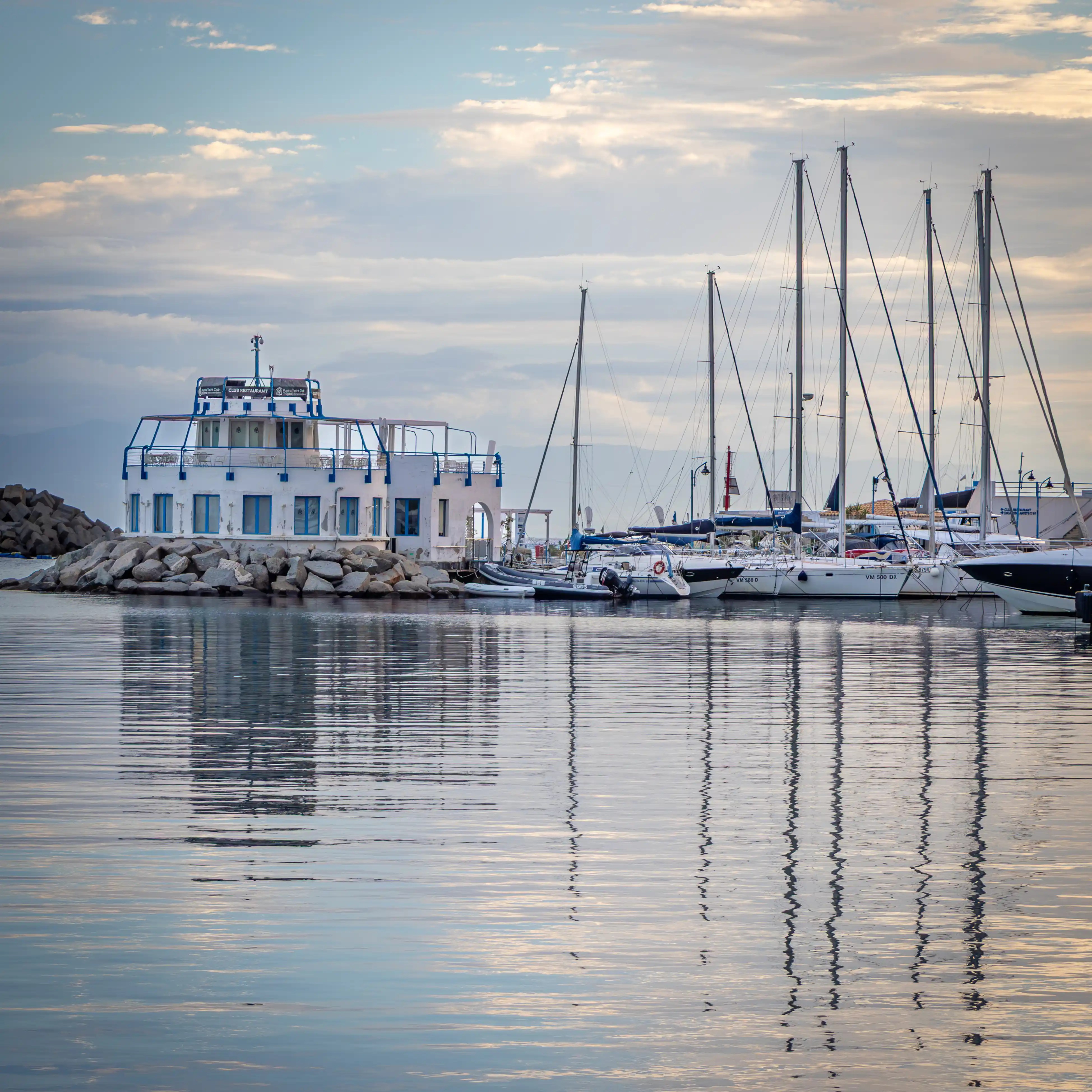 A white building on a breakwater with sailboats nearby and reflections in calm water.