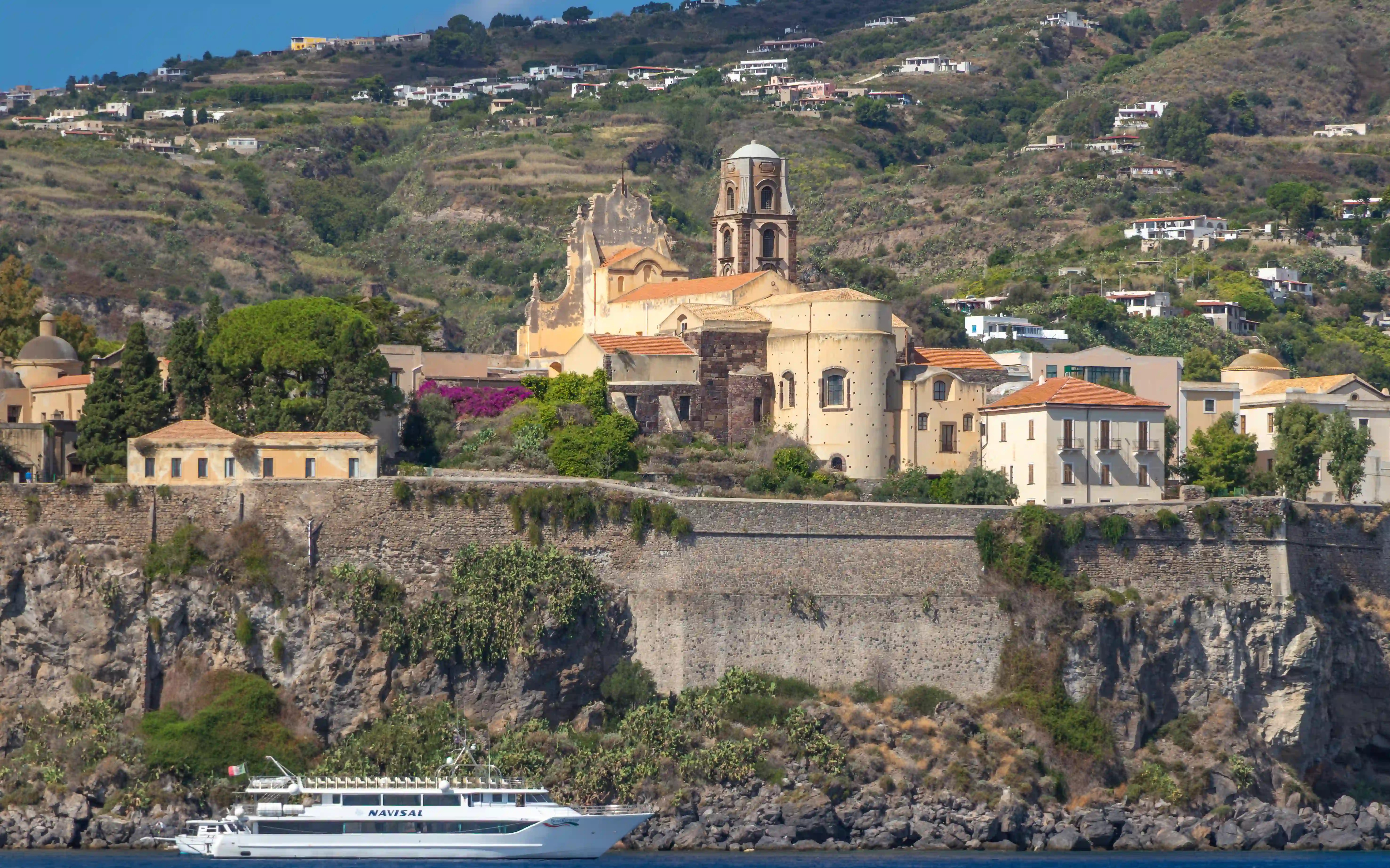 Church buildings and a bell tower on a stone wall above rocky cliffs with a boat passing below.
