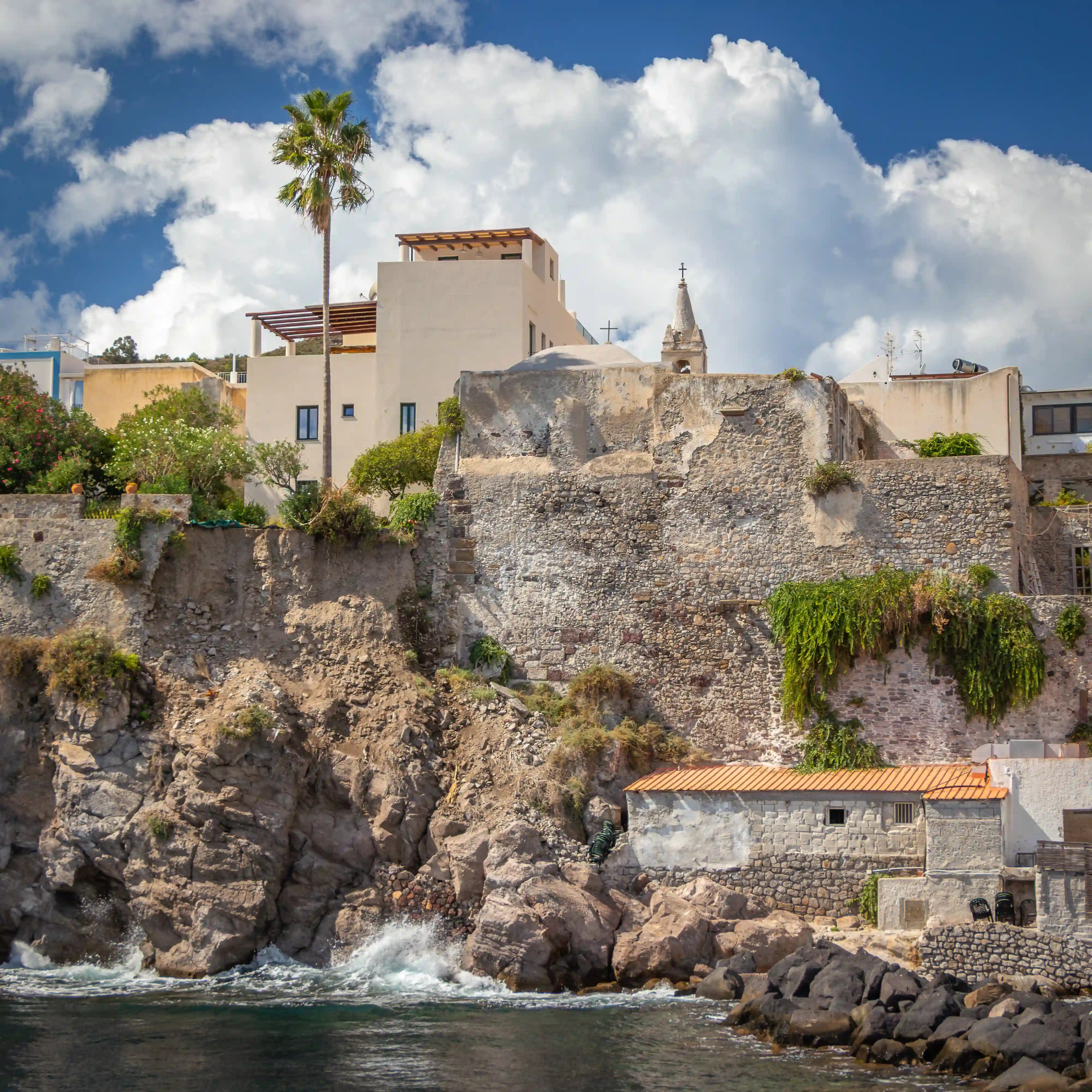 Stone wall and buildings on a rocky cliff above the water with a tall palm tree and cloudy sky.
