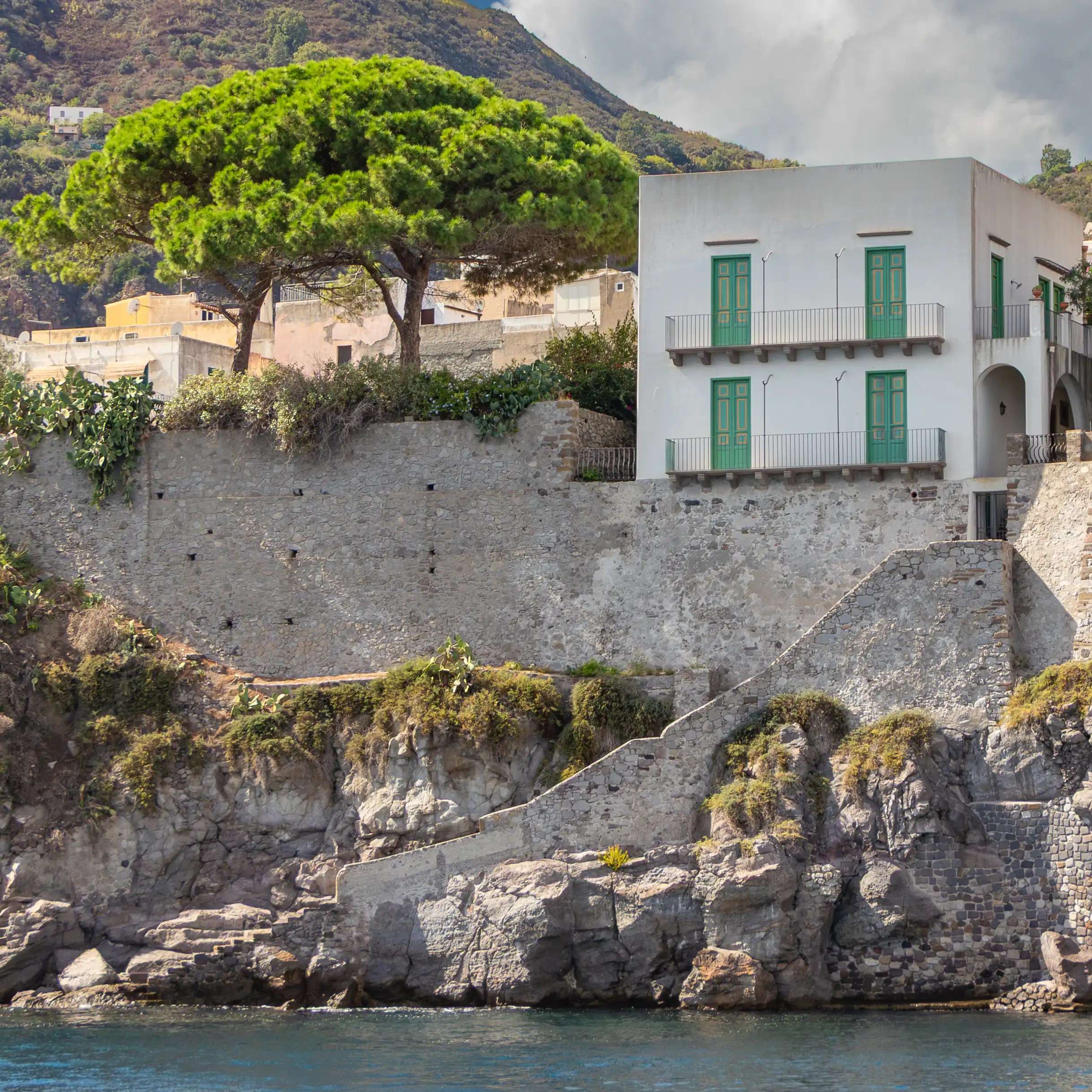 White building with green shutters and balconies above a stone wall and staircase along the rocky shoreline.