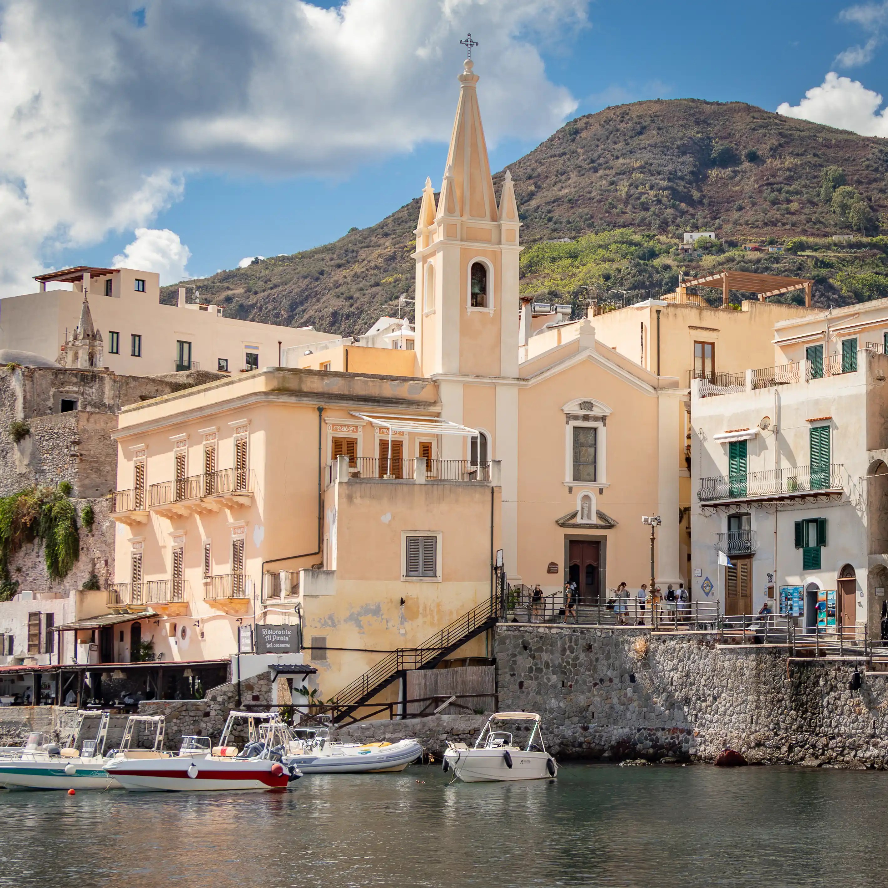 Church with a tall spire beside pastel buildings along a waterfront with small boats in the harbor.