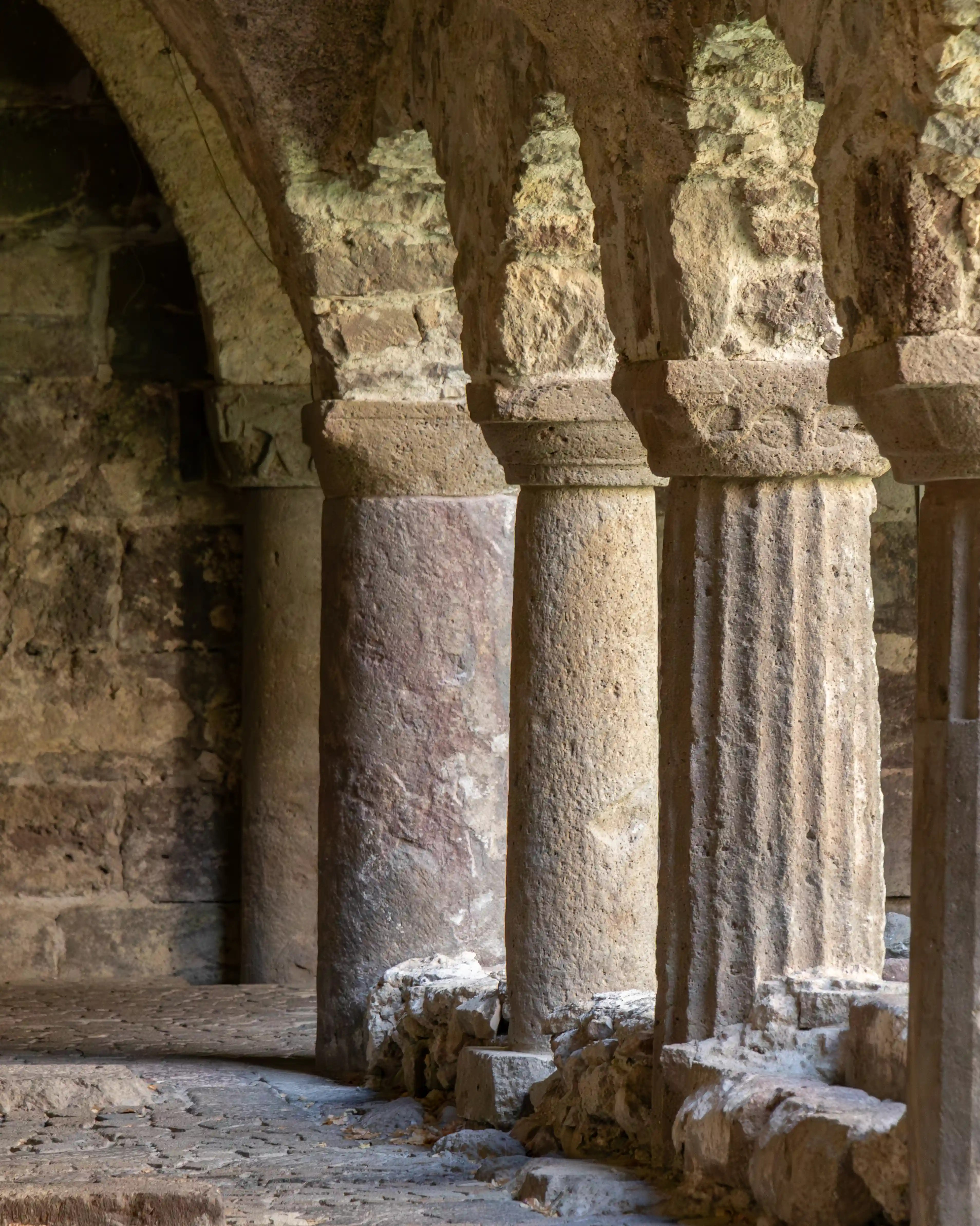 Close view of stone columns and arches in a dimly lit walkway with textured surfaces and worn edges.