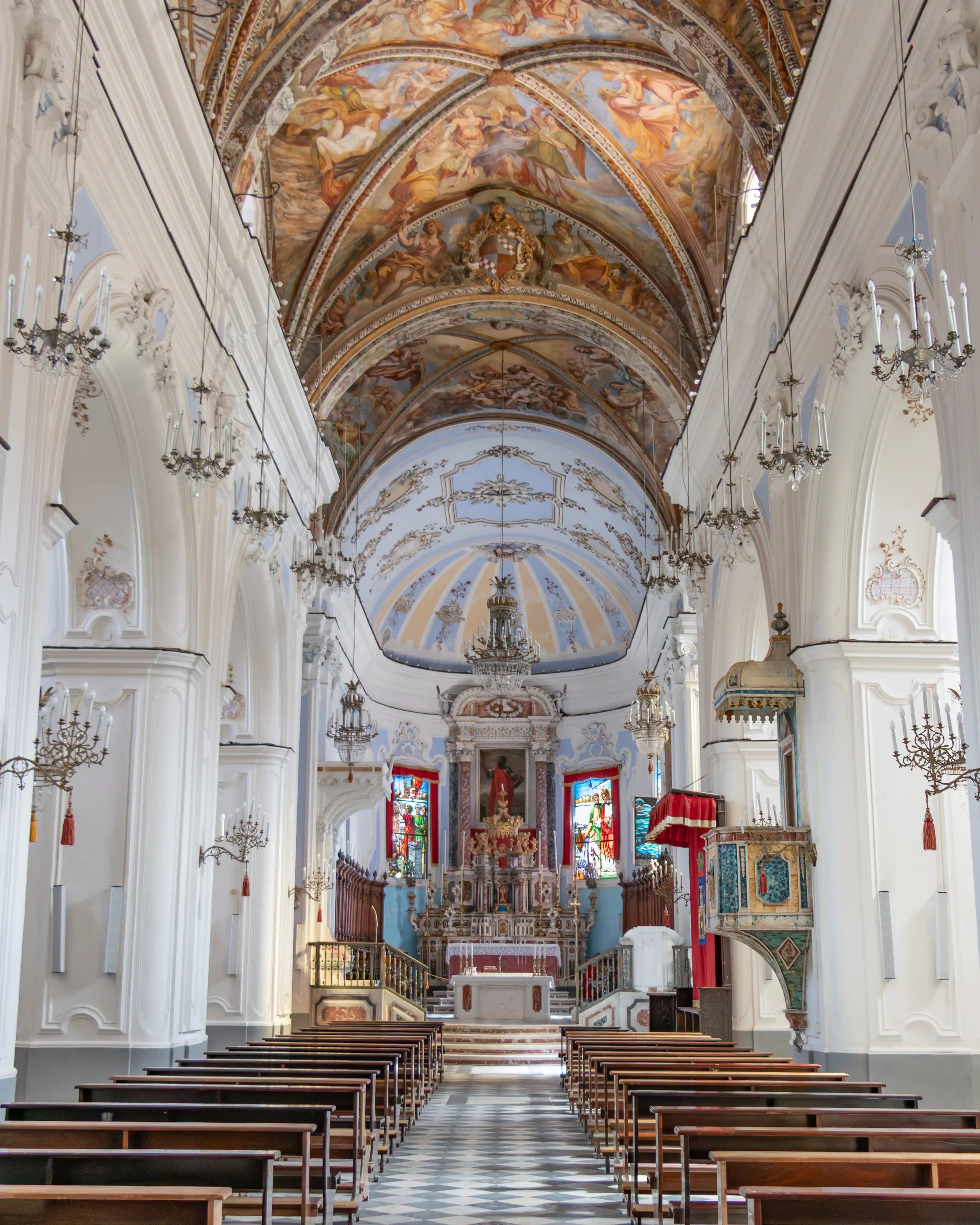 Interior of a church with rows of wooden pews, chandeliers, and a painted vaulted ceiling above an altar.