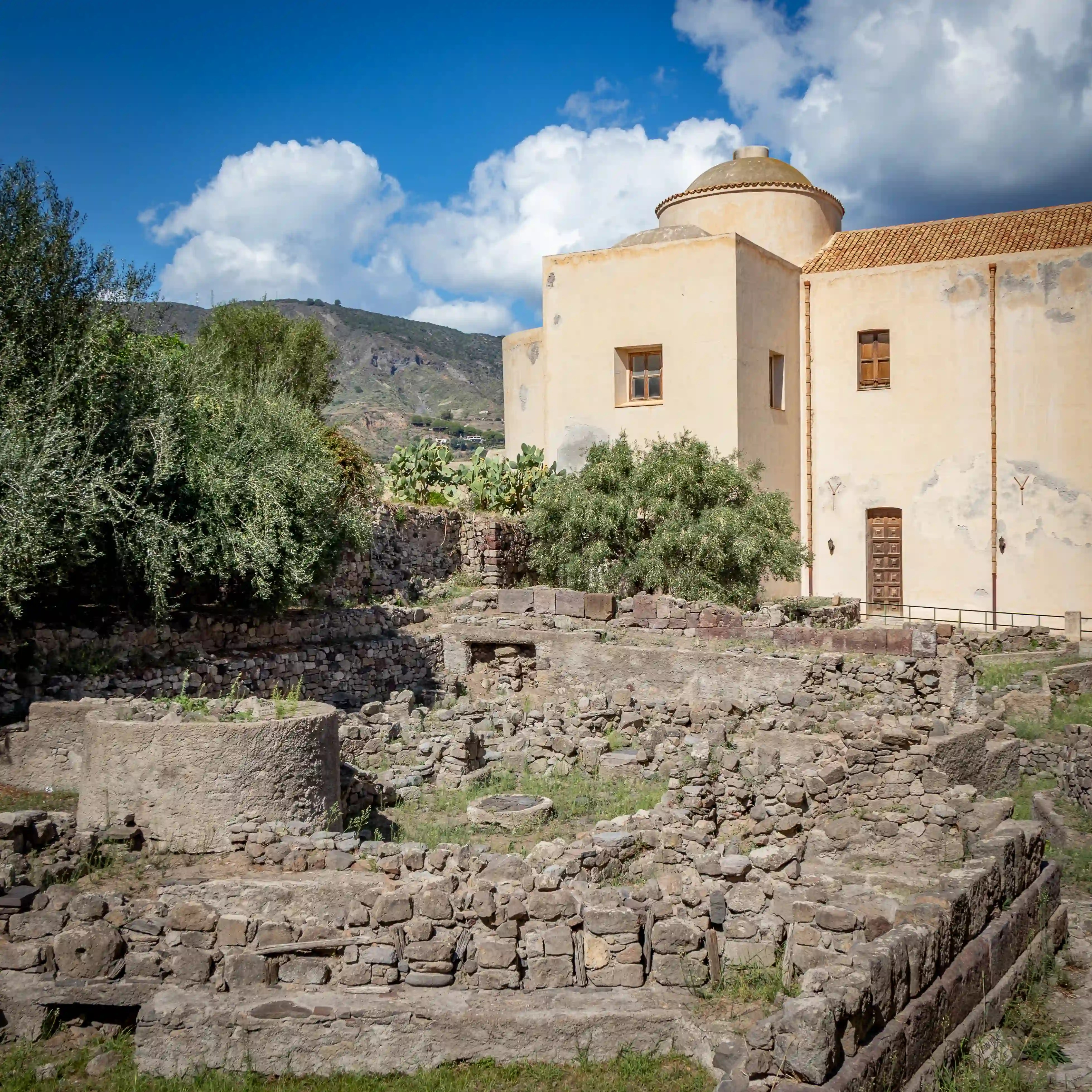Stone ruins and excavation walls in an archaeological site below a pale yellow building with a dome in Lipari.