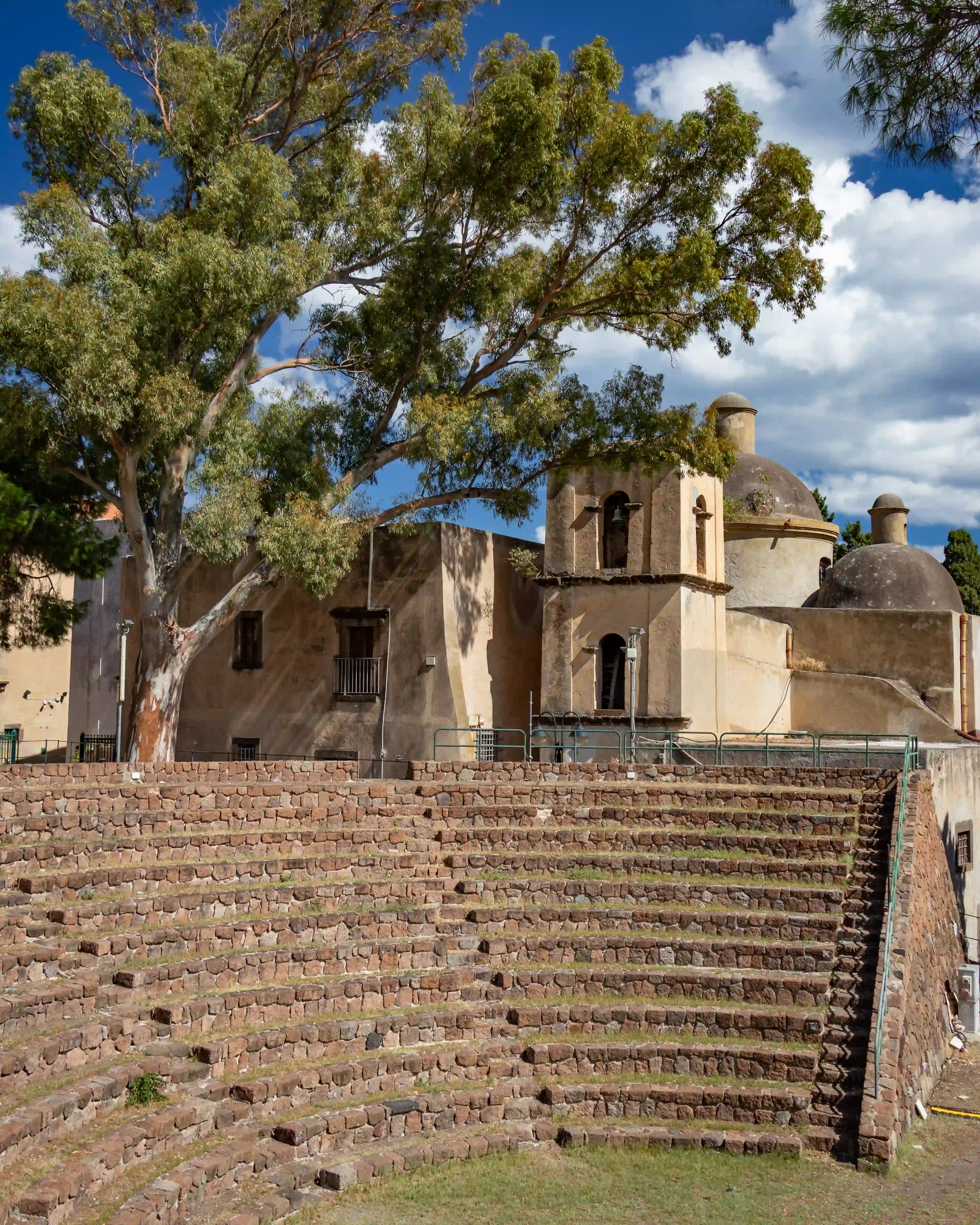 Curved stone seating forming a small amphitheater in front of a light-colored building with domes and a bell tower.