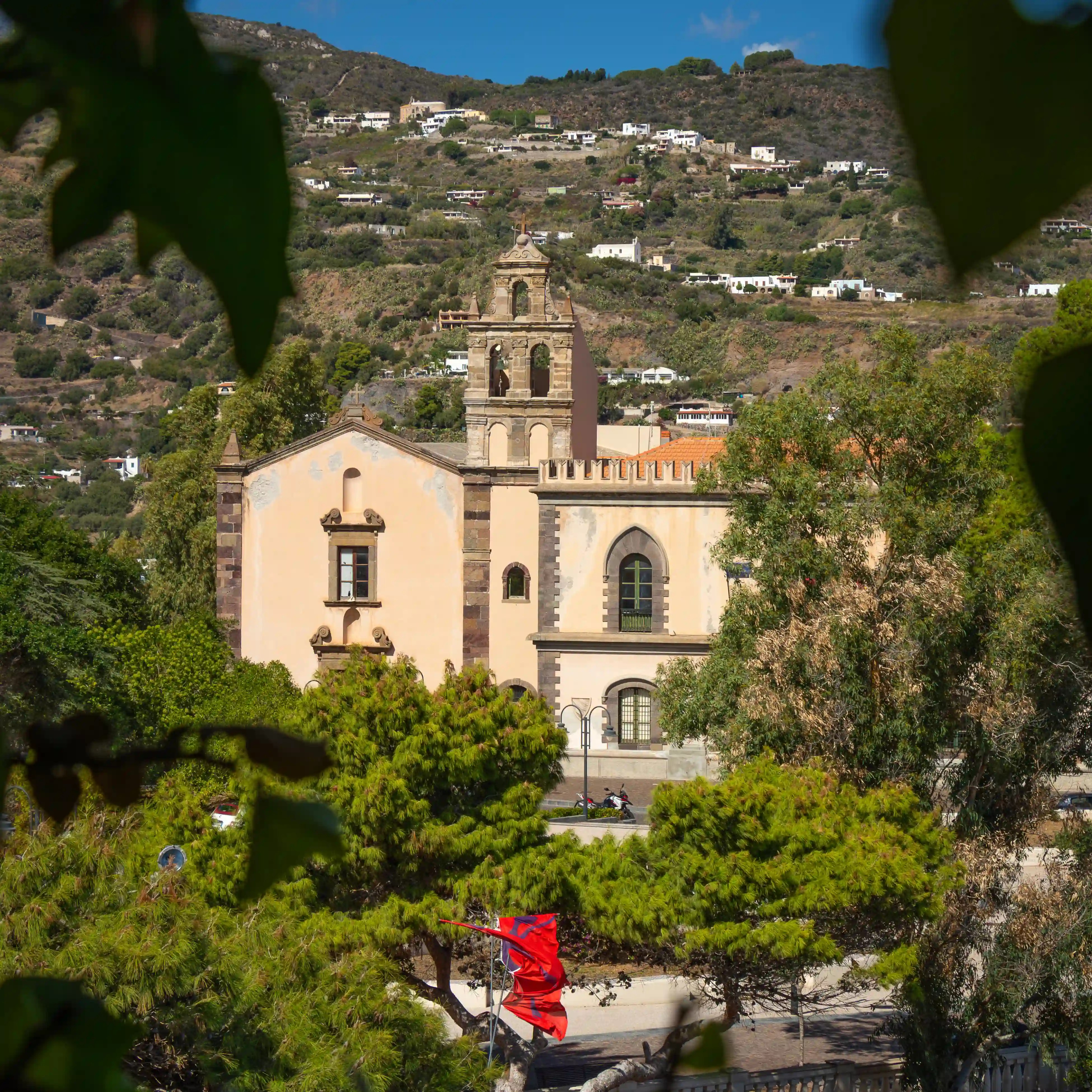 A church with a bell tower framed by tree branches, with houses scattered across a hillside in the background.