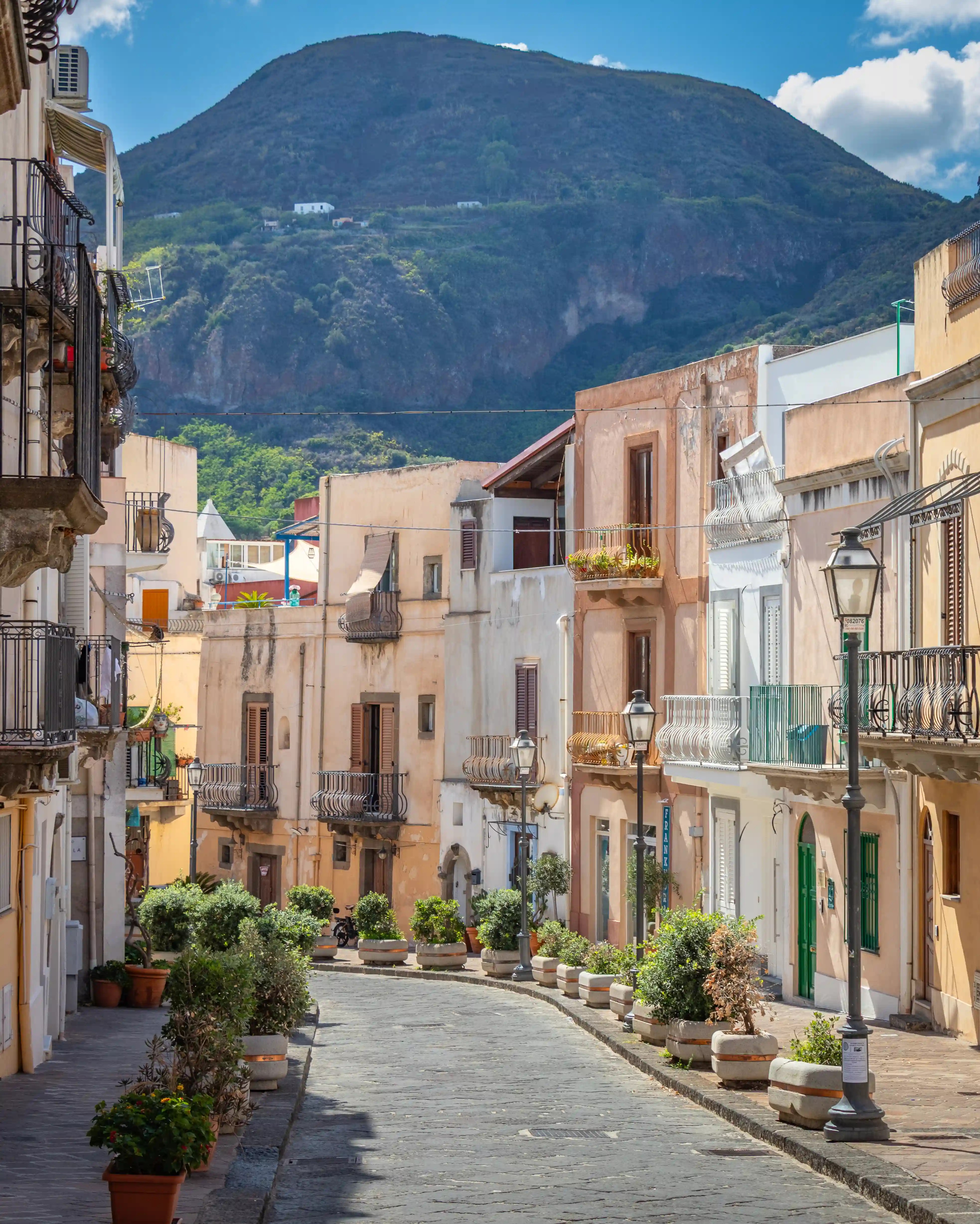 A quiet street lined with pastel buildings, balconies, and potted plants, with a large mountain rising in the background.