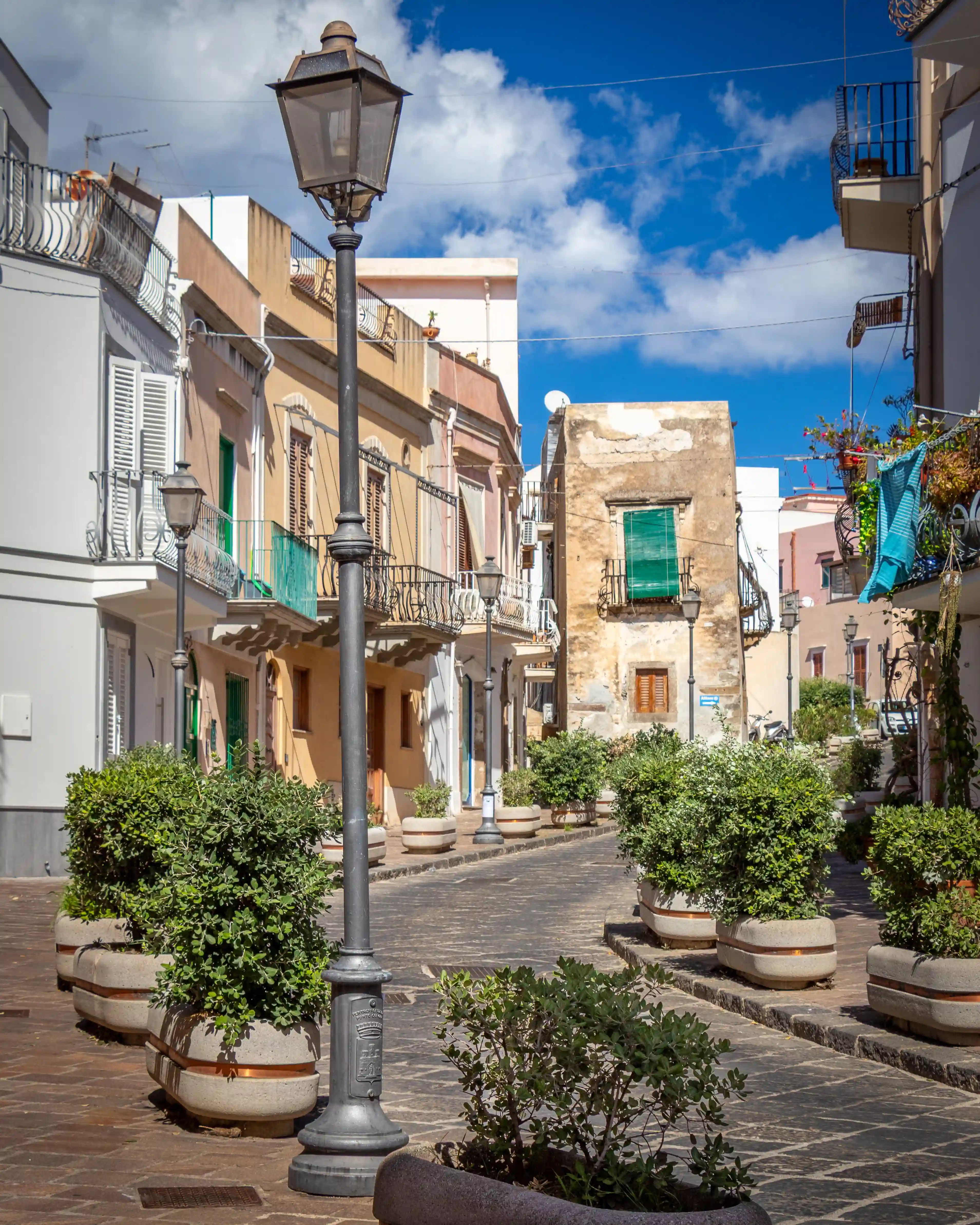 A narrow street with a central lamppost, surrounded by low buildings, balconies, and large potted plants.