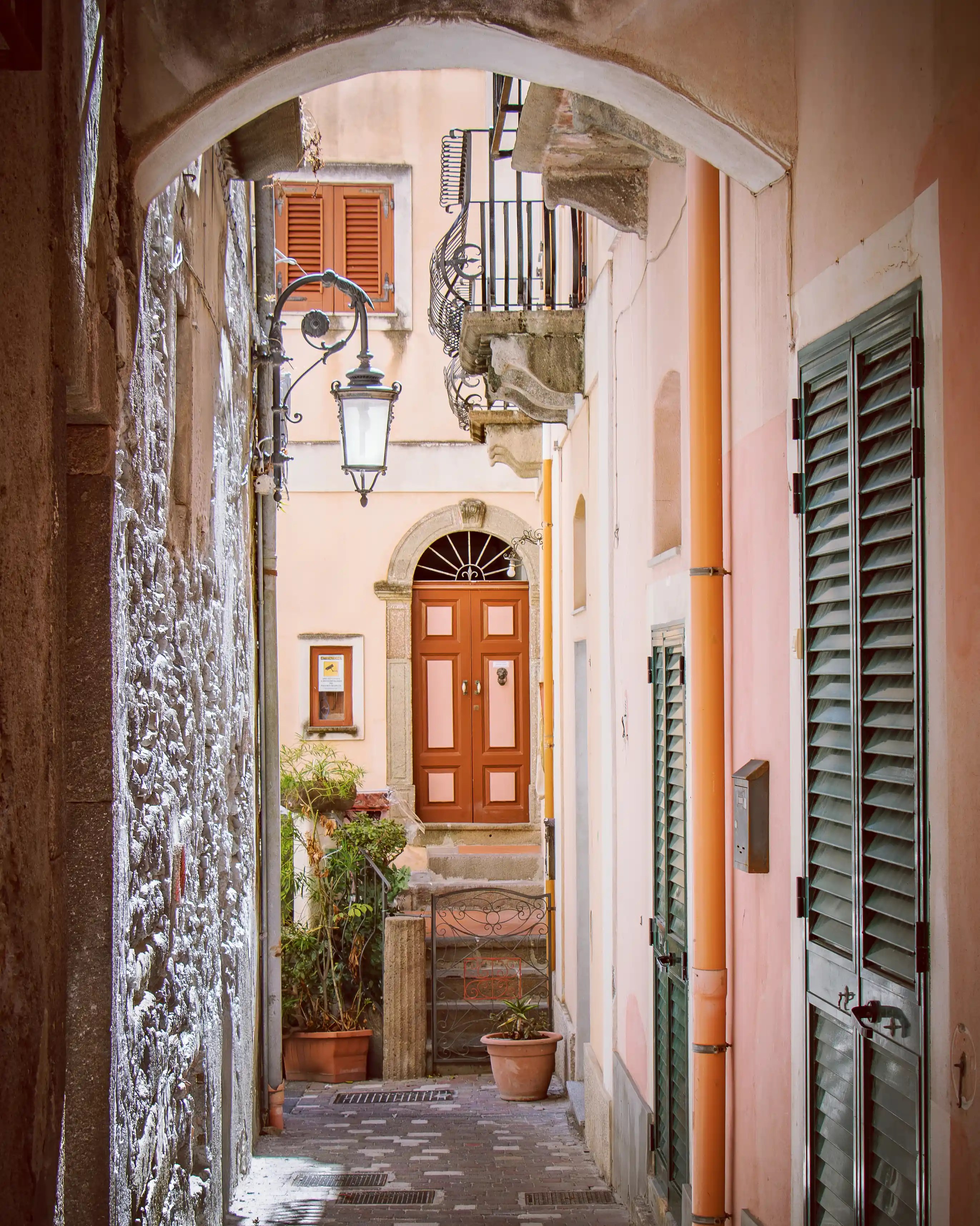 A narrow alleyway with an arched passage, leading to a red door at the end and lined with shutters and balconies.