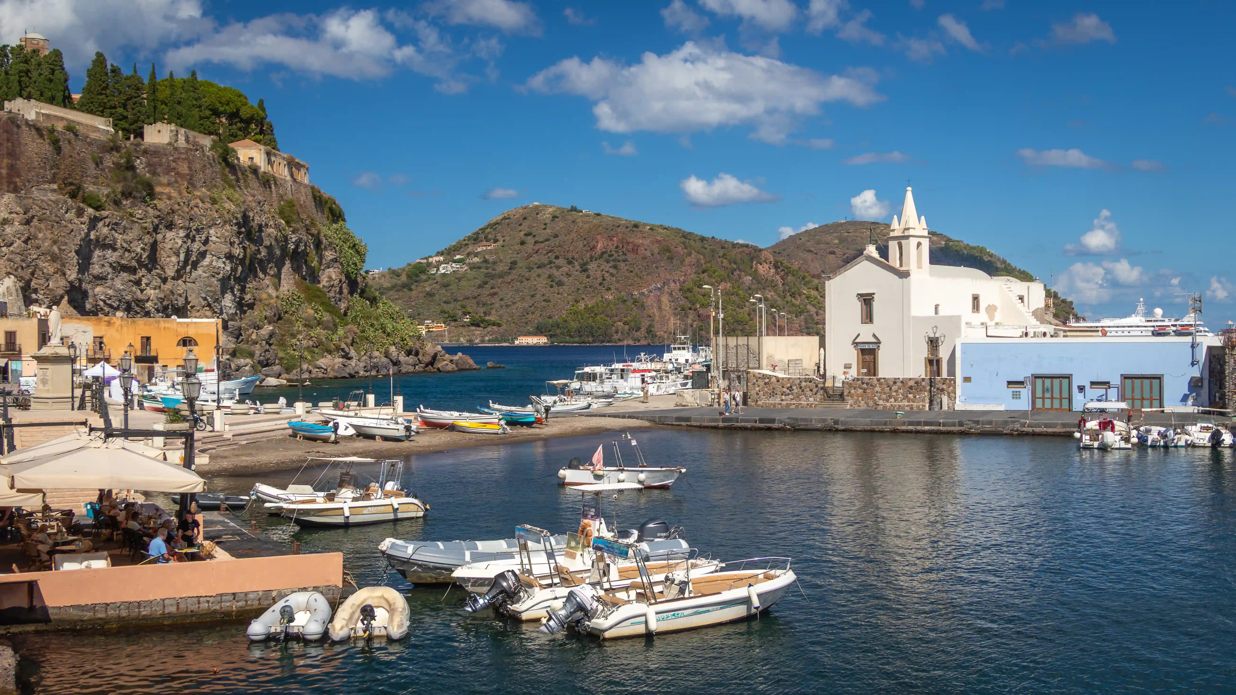 A harbor with small boats, a white church on the waterfront, and rocky cliffs rising behind the town.
