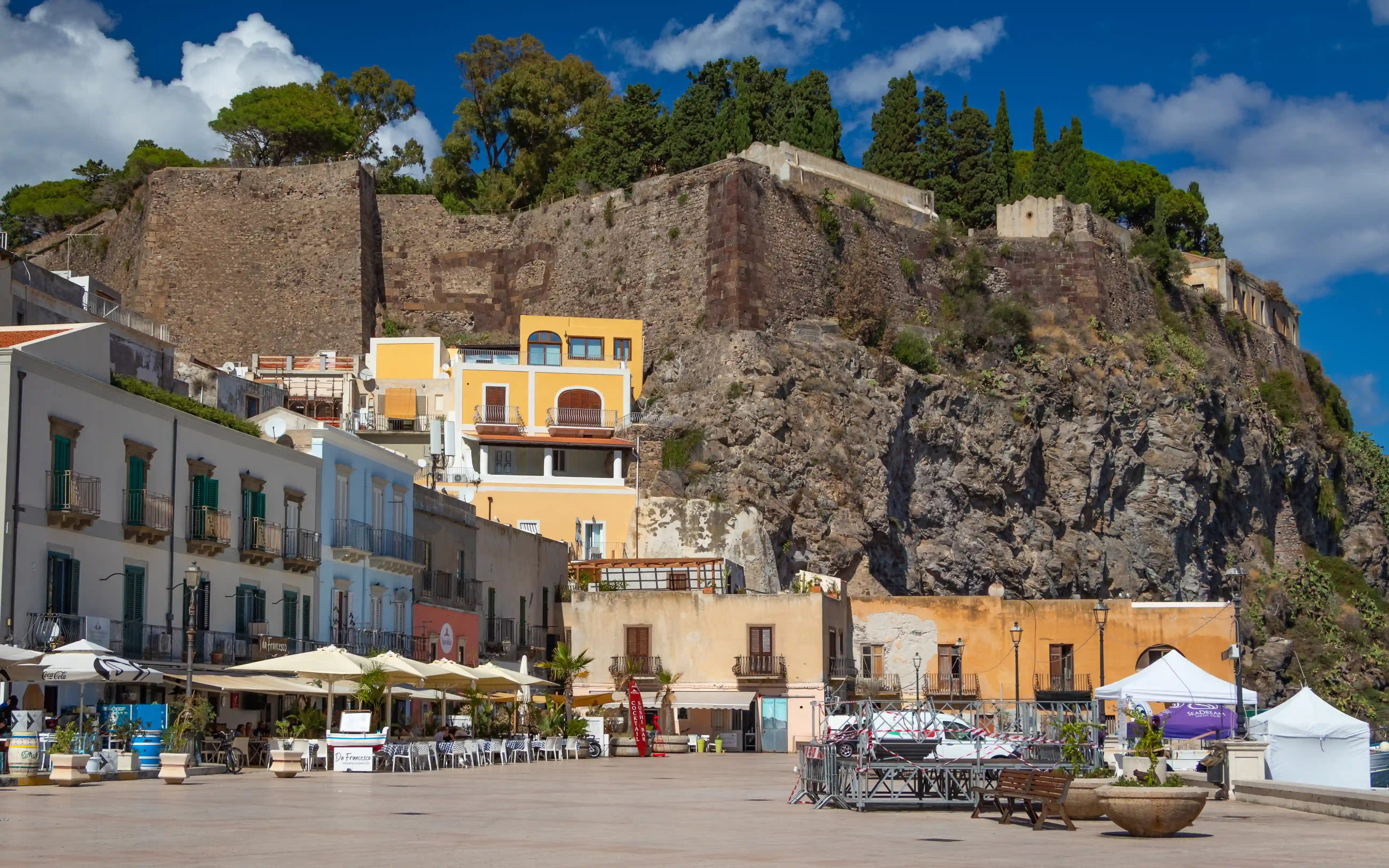 A large stone fortress wall on top of a rocky cliff above a square lined with buildings and outdoor seating.
