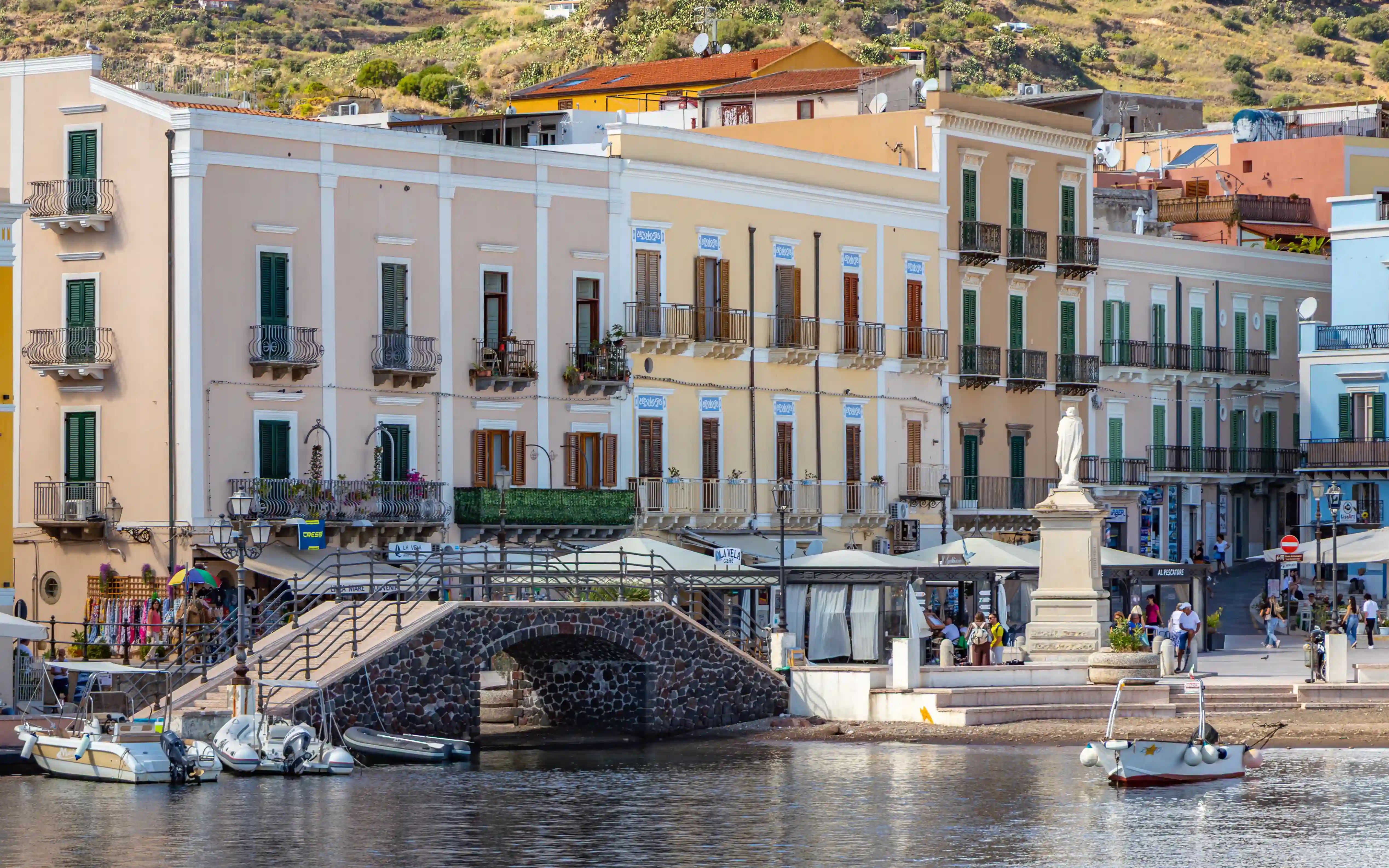 Pastel-colored buildings along a waterfront with a small bridge, a statue on a pedestal, and boats in the water.