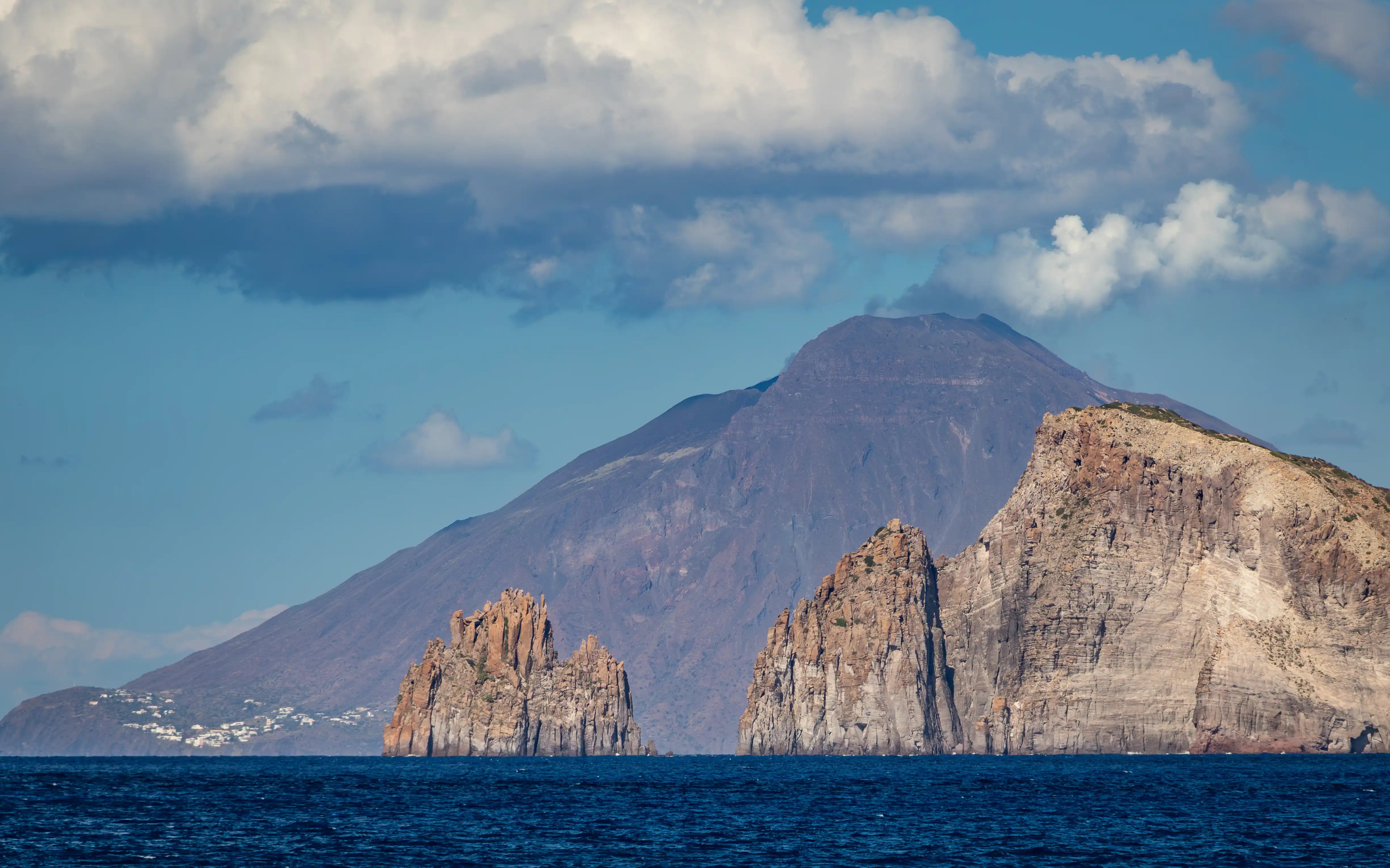 Rock formations rising from the sea with a large volcanic island in the background under a partly cloudy sky.