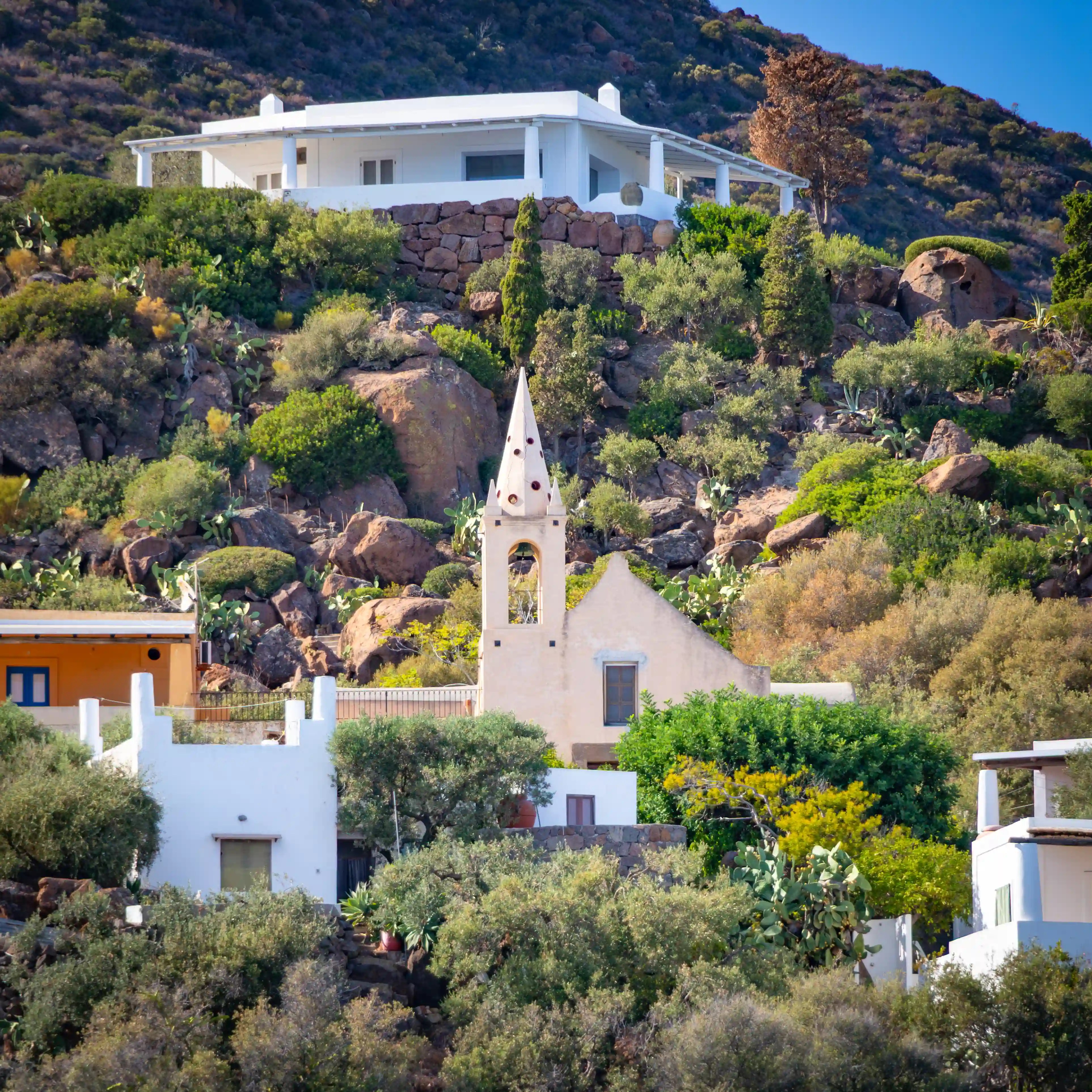 A small light-colored chapel with a narrow bell tower surrounded by houses and vegetation on a rocky hillside.