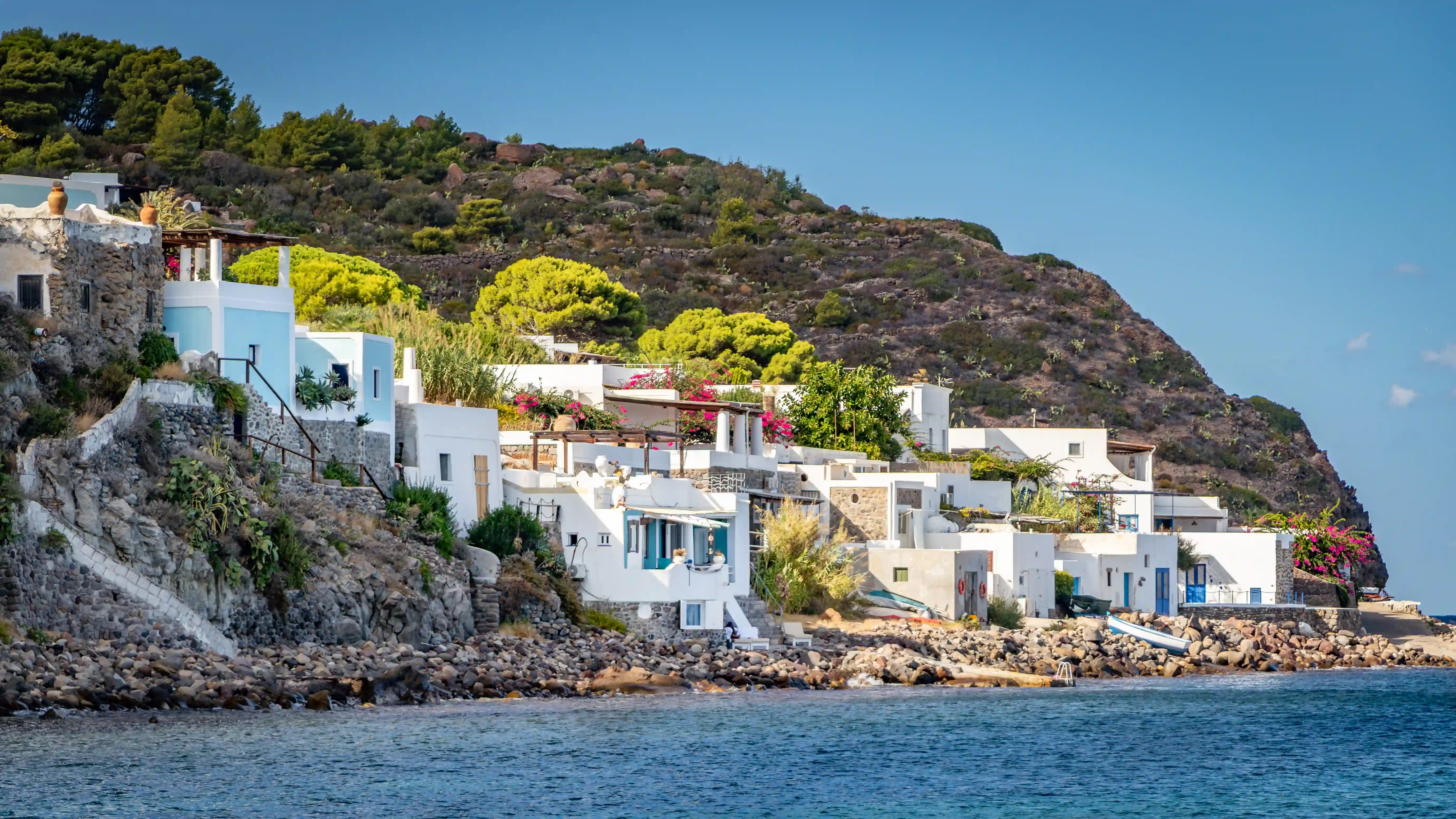White houses with terraces and greenery line a rocky shoreline beside calm blue water.