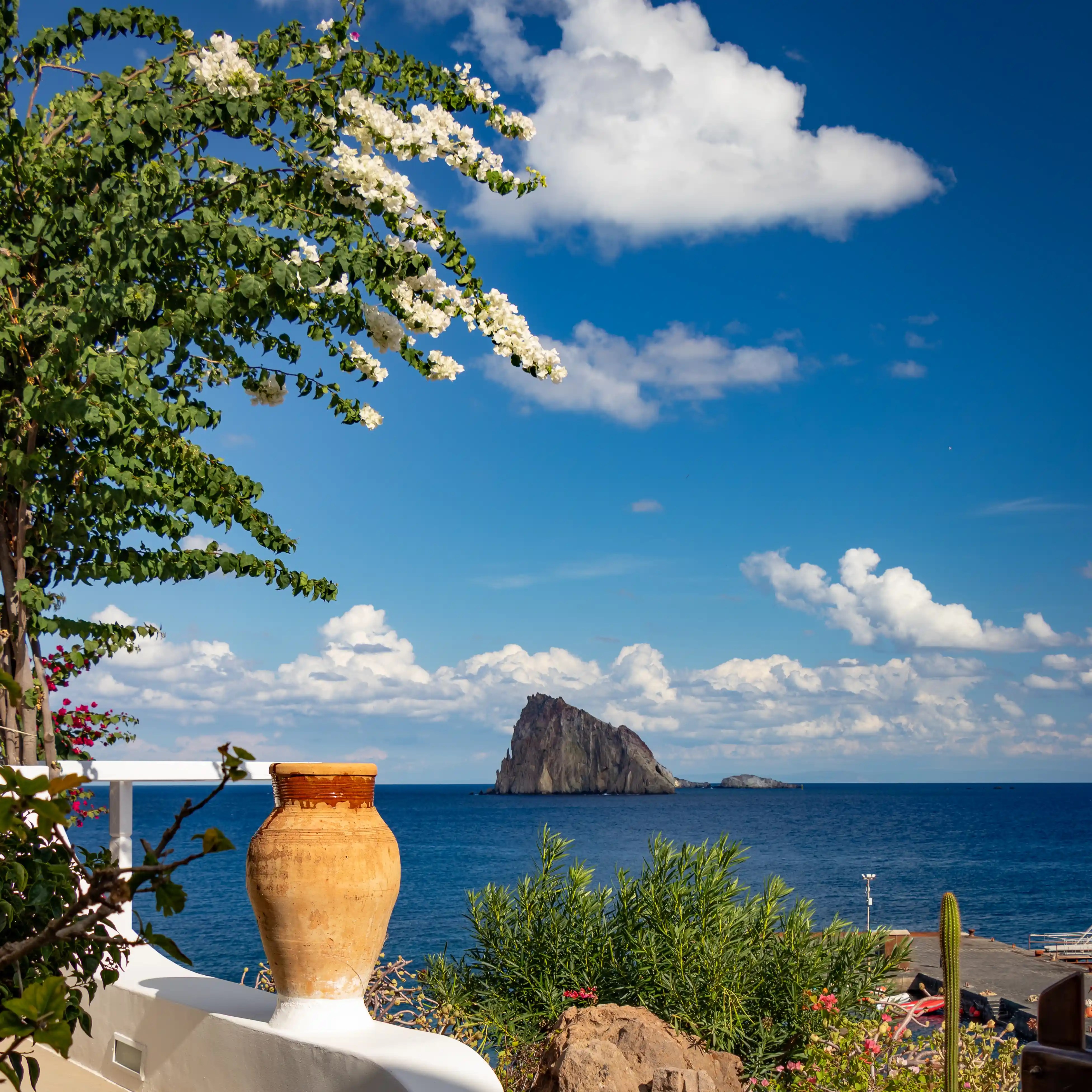 A large clay pot and flowering branches frame a view of a rocky islet in the sea under a blue sky with clouds.