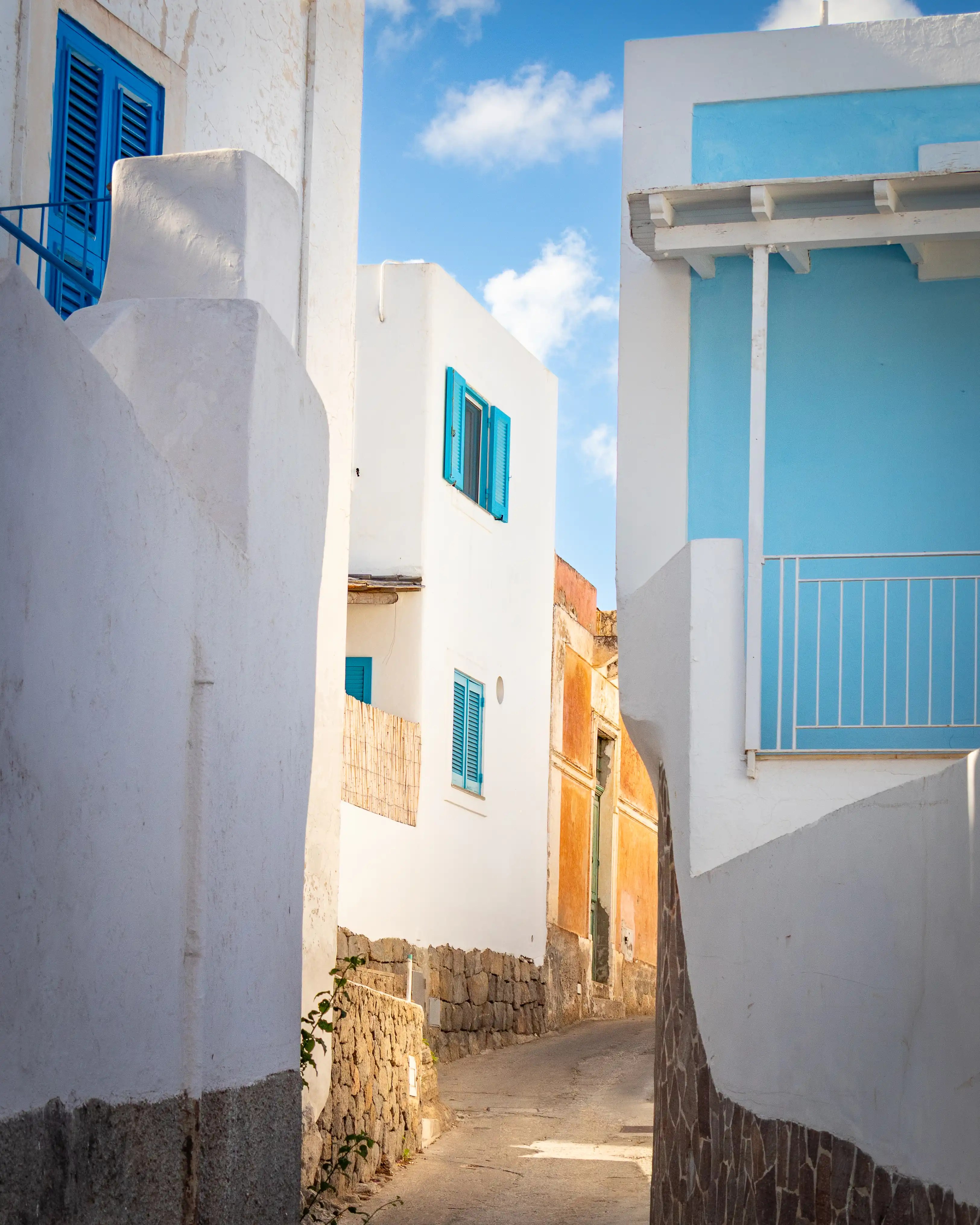 A narrow paved alley between white buildings with blue shutters and a small balcony.