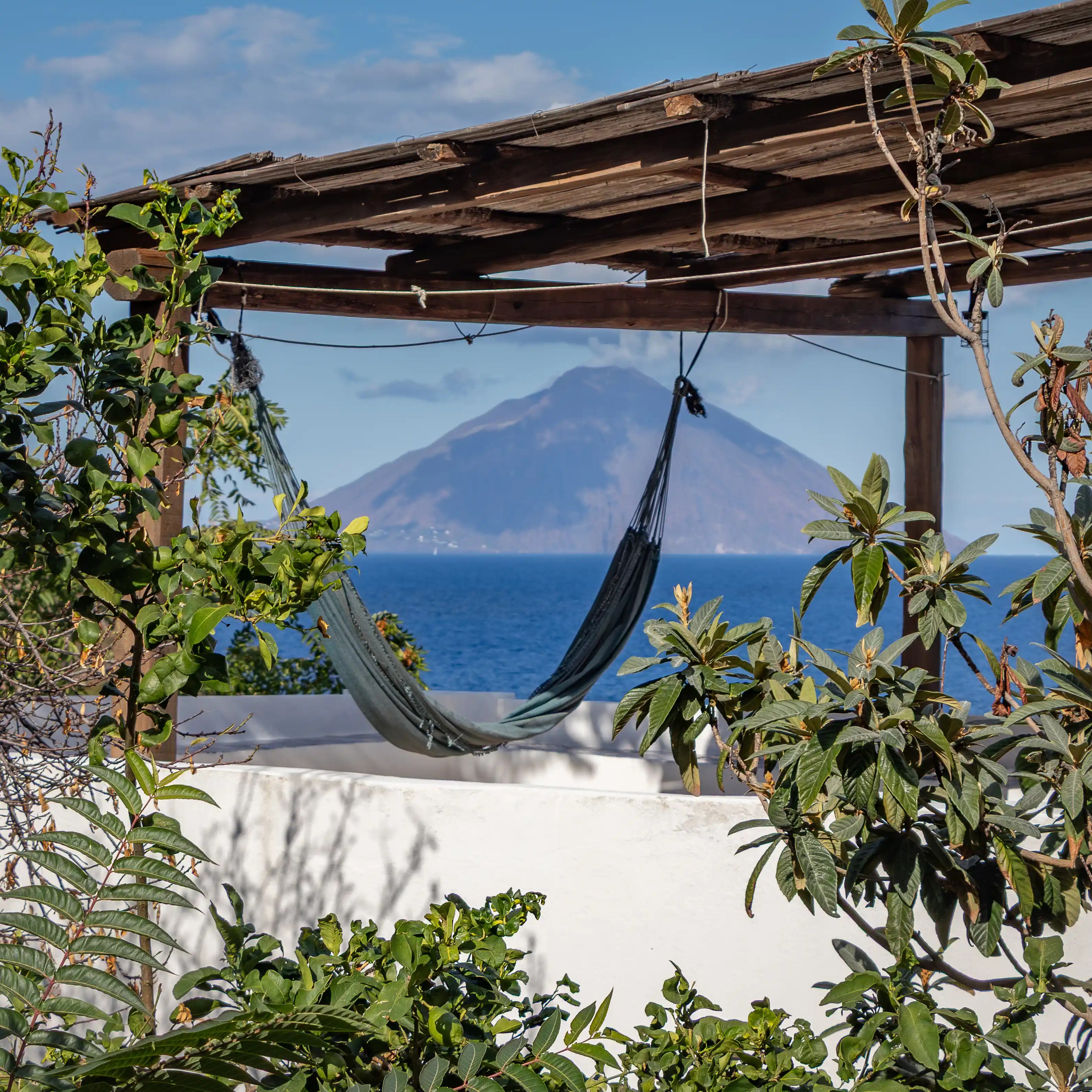 A hammock hangs under a wooden pergola overlooking the sea with a volcanic island in the distance.