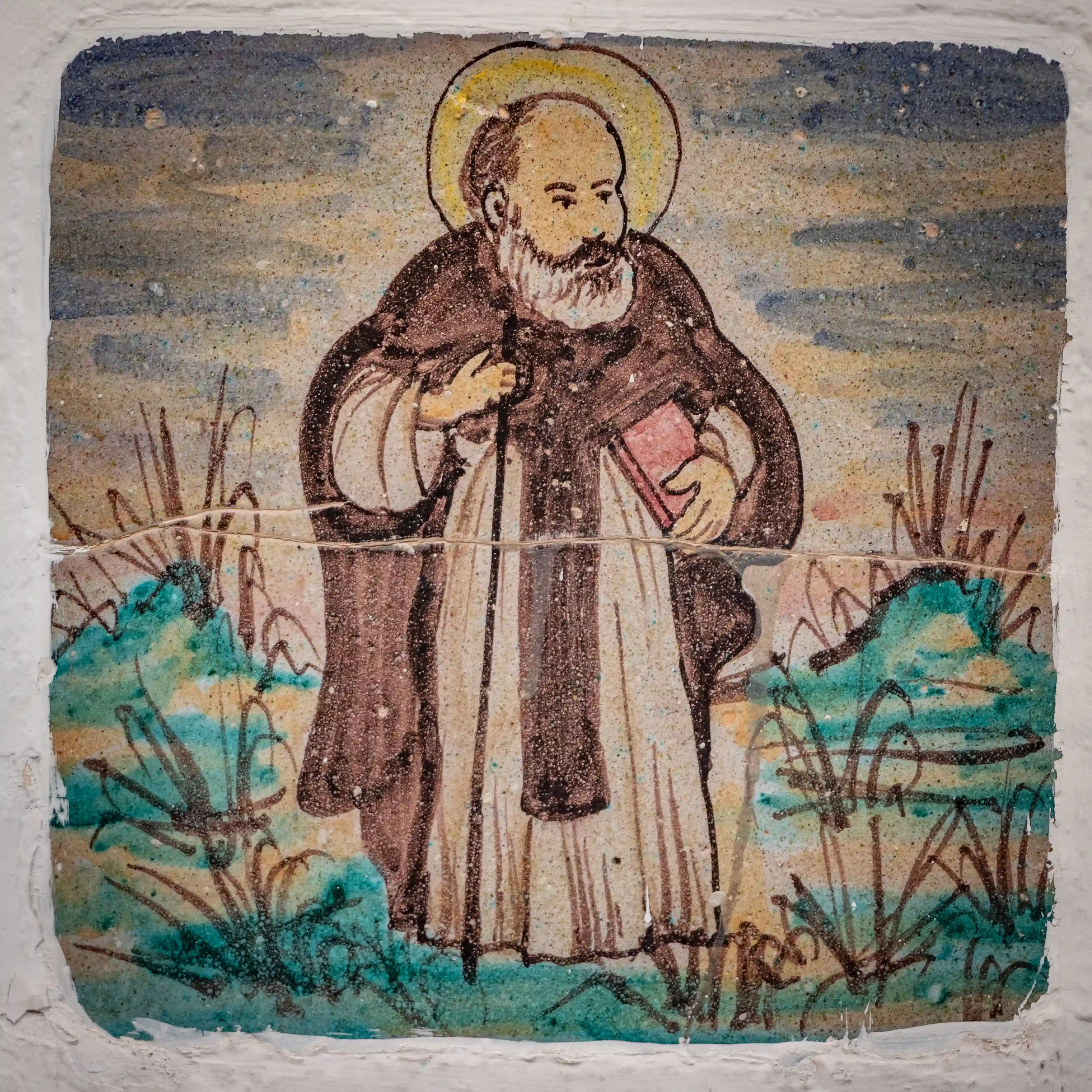 A ceramic tile depicting a bearded saint holding a book and staff with a halo around his head.