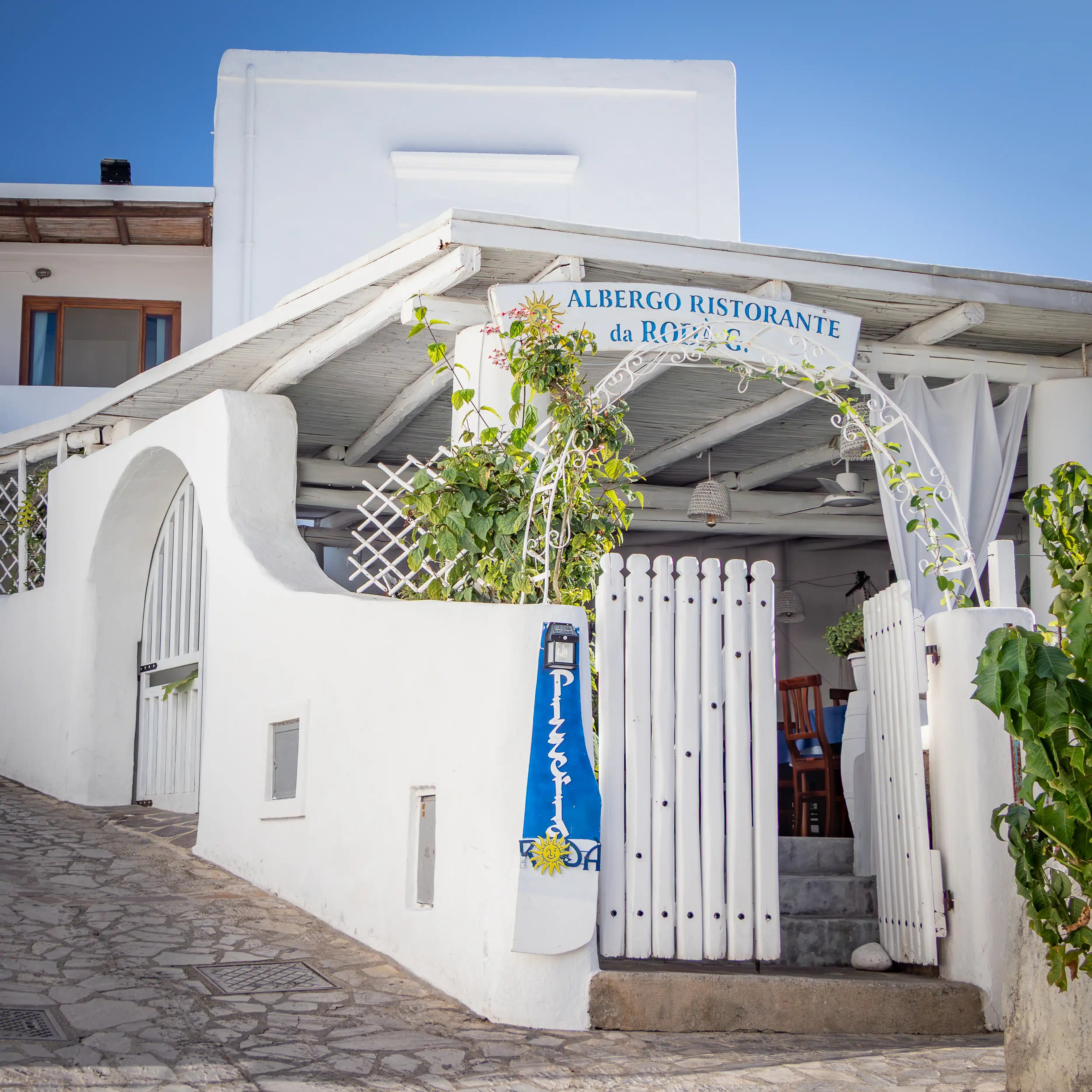 A white building entrance with a sign reading “Albergo Ristorante da Rodà G.” above a gate and pergola.