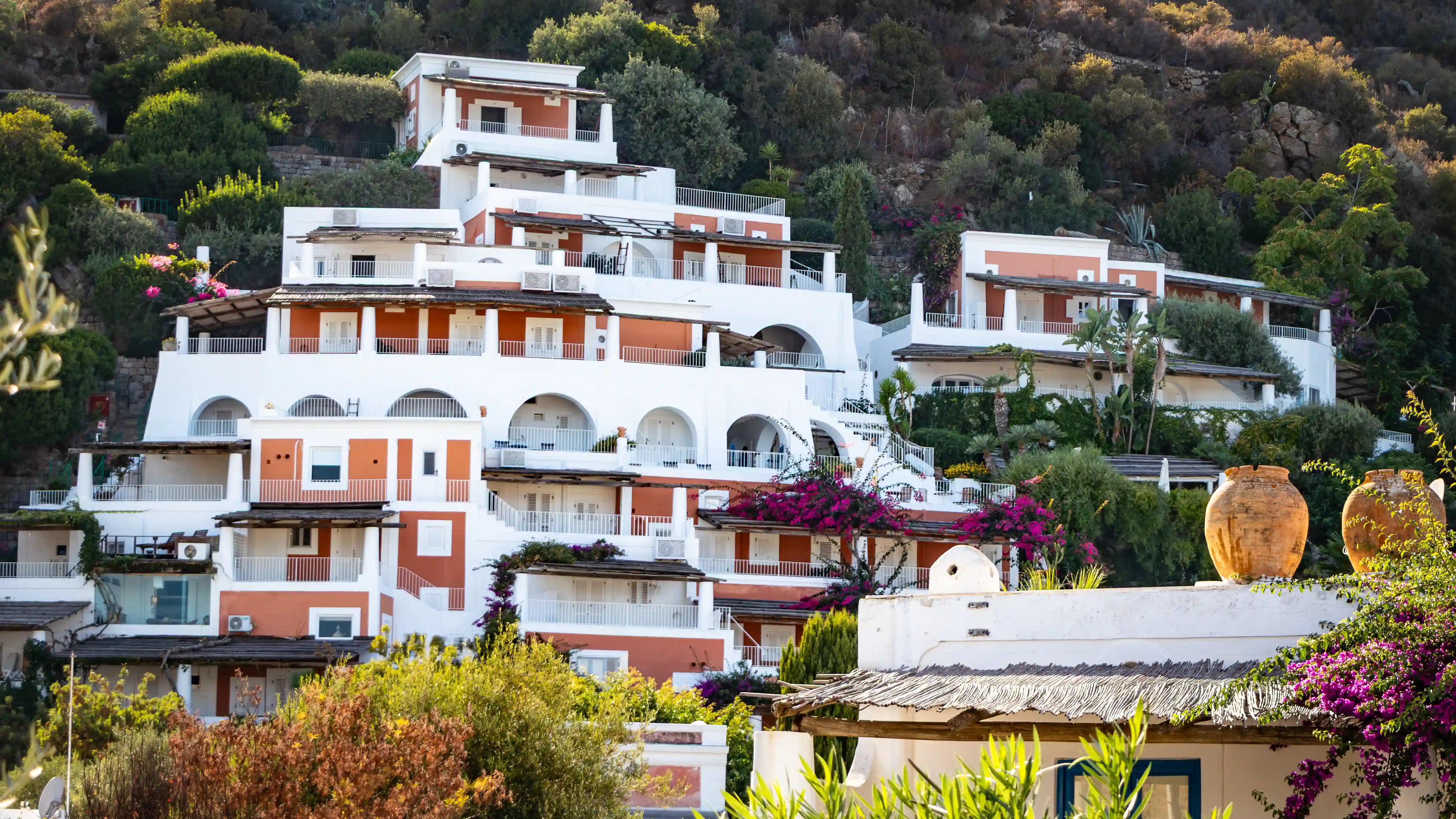 White and peach-colored buildings with terraces and arches arranged on a hillside with vegetation.