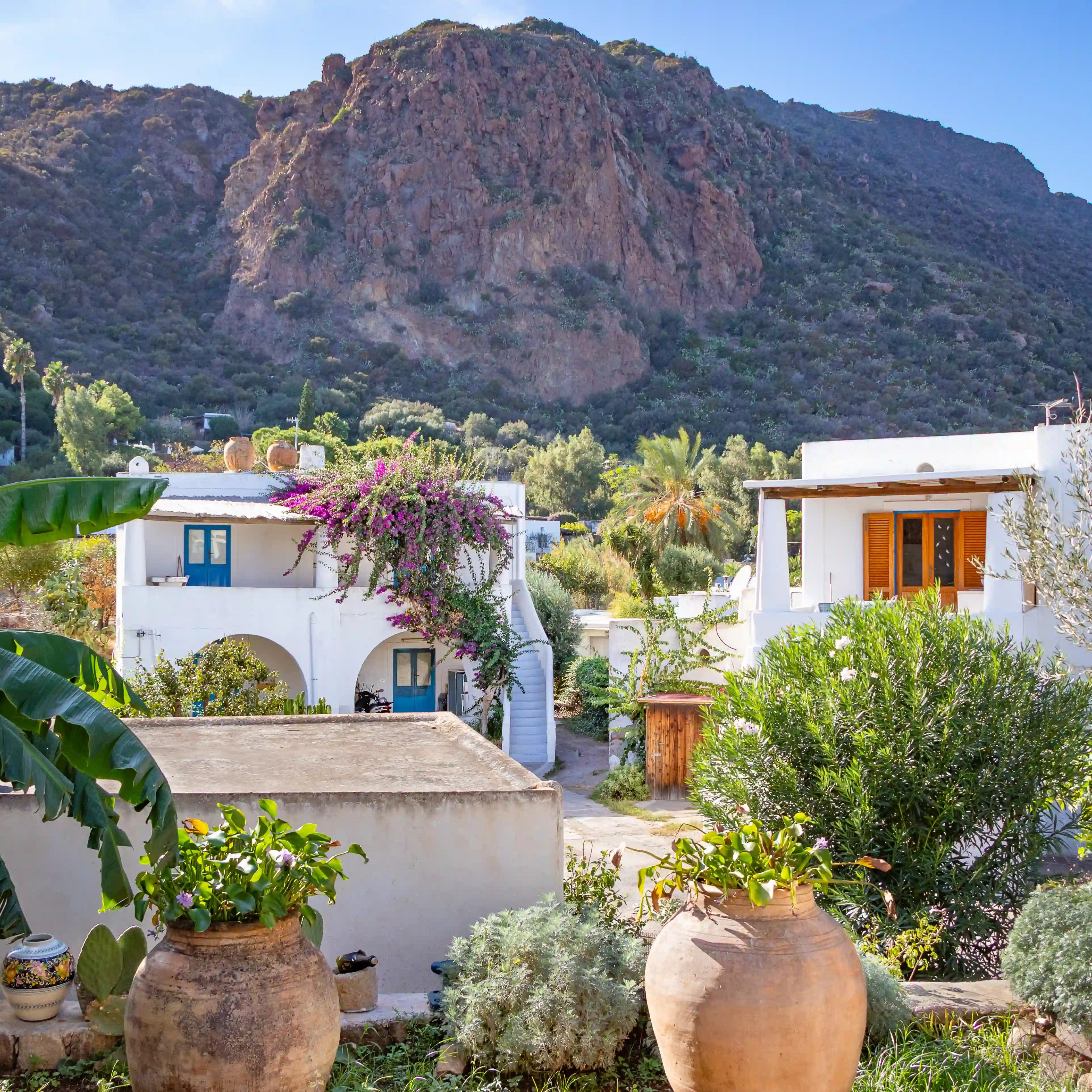 White houses with plants and clay pots in the foreground and a rocky hill rising behind them.